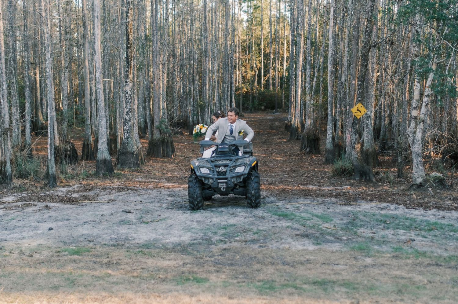 Bride and groom ride an ATV through a wooded forest path on their wedding day.