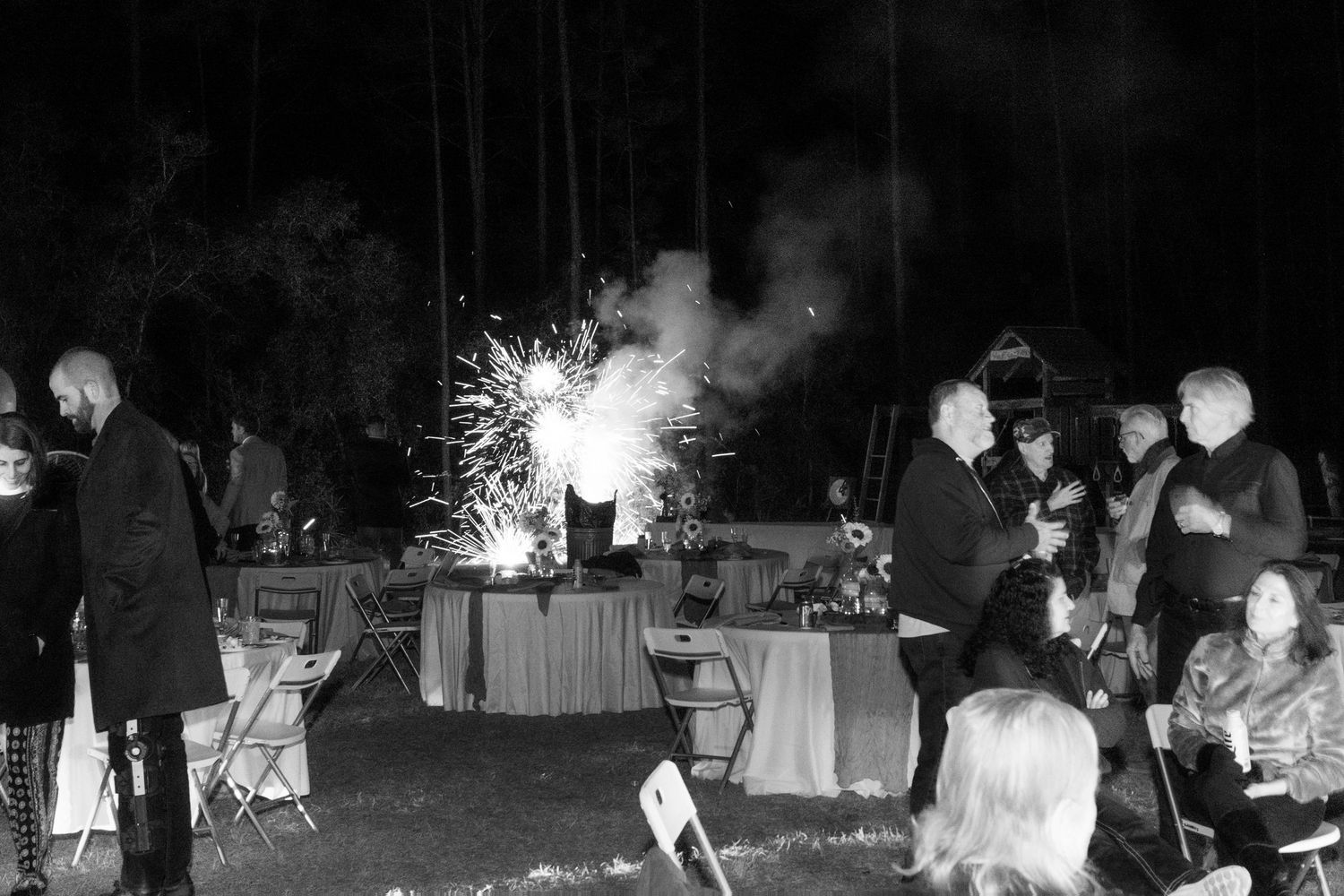 Wedding guests gather outdoors at night around tables as sparklers light up the dark celebration.
