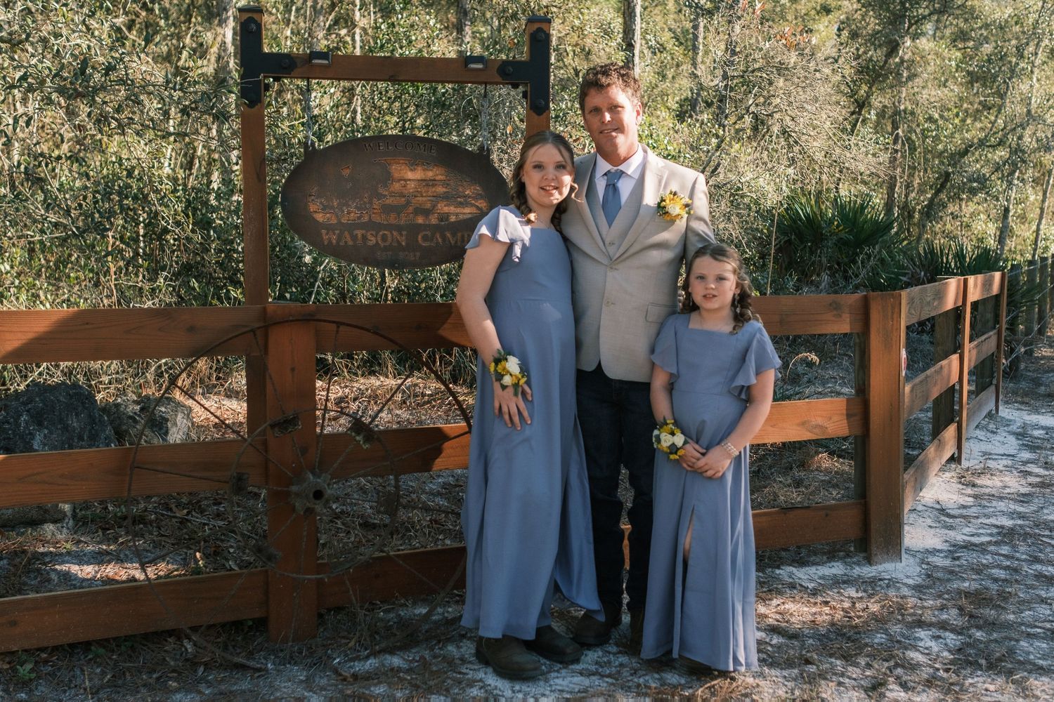 Man with two girls in blue dresses posing by wooden sign and fence at wedding venue.