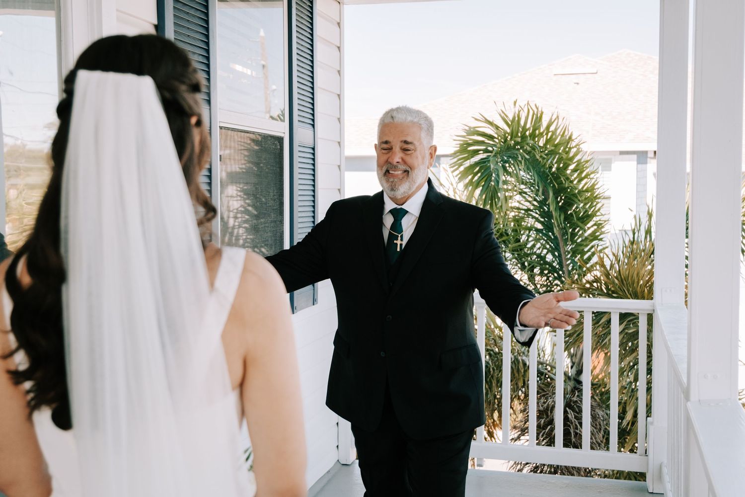 A person in a black suit reacts with joy upon seeing a bride in her white wedding dress on a porch.