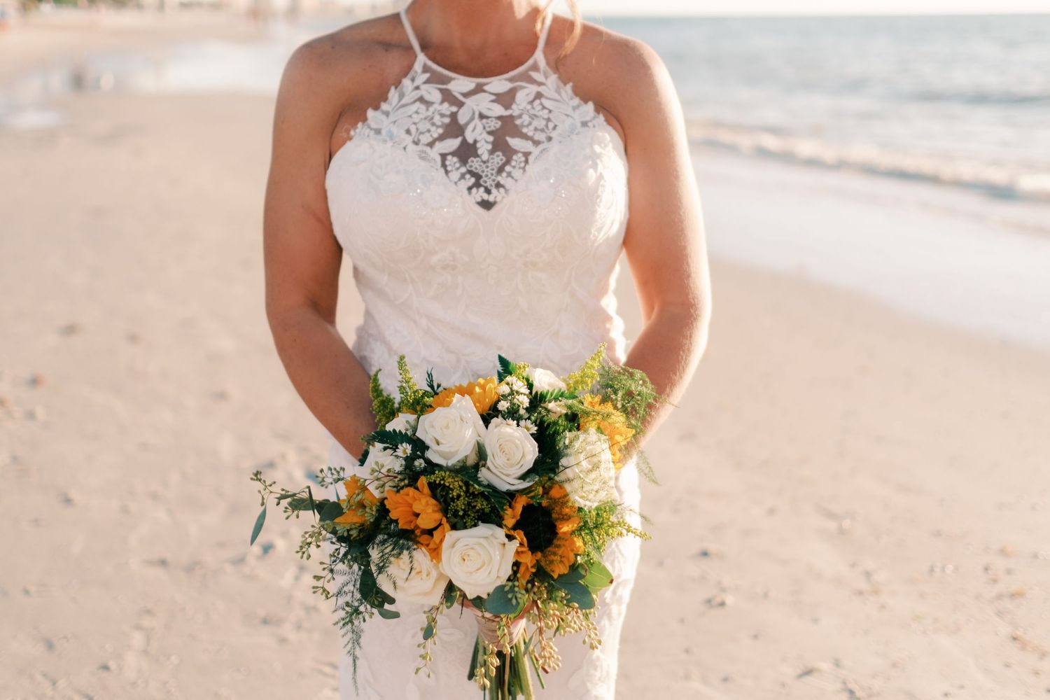 Bride in halter lace wedding gown holds vibrant bouquet of white and orange flowers on sandy beach.