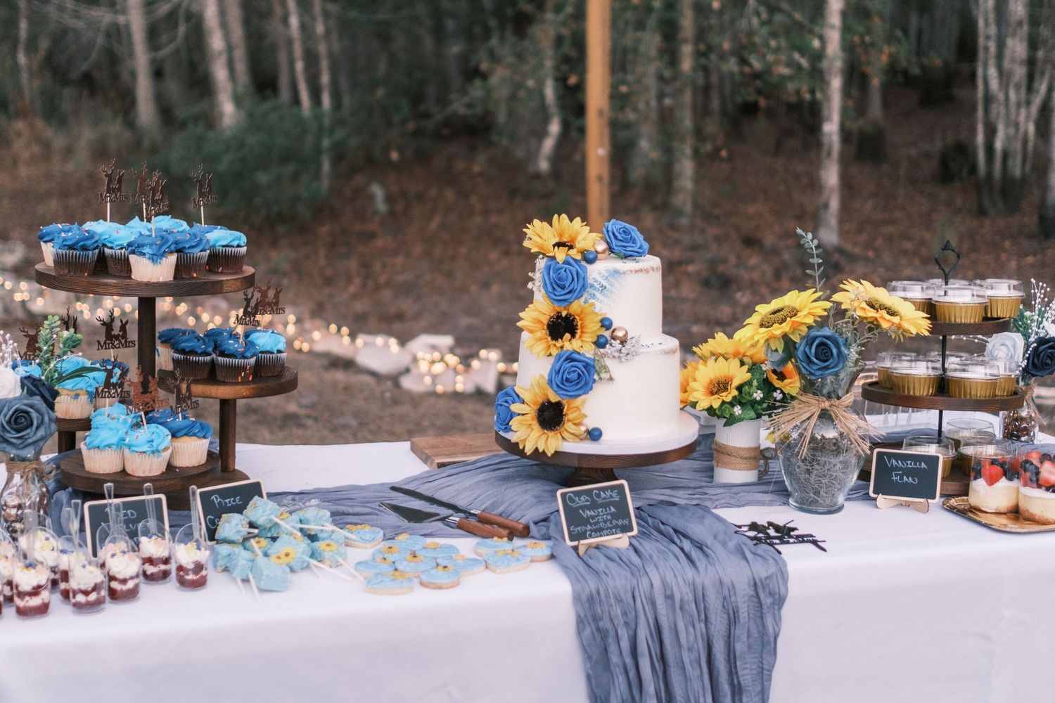 Wedding dessert table with white cake, blue cupcakes, sunflowers, and assorted sweets outdoors.