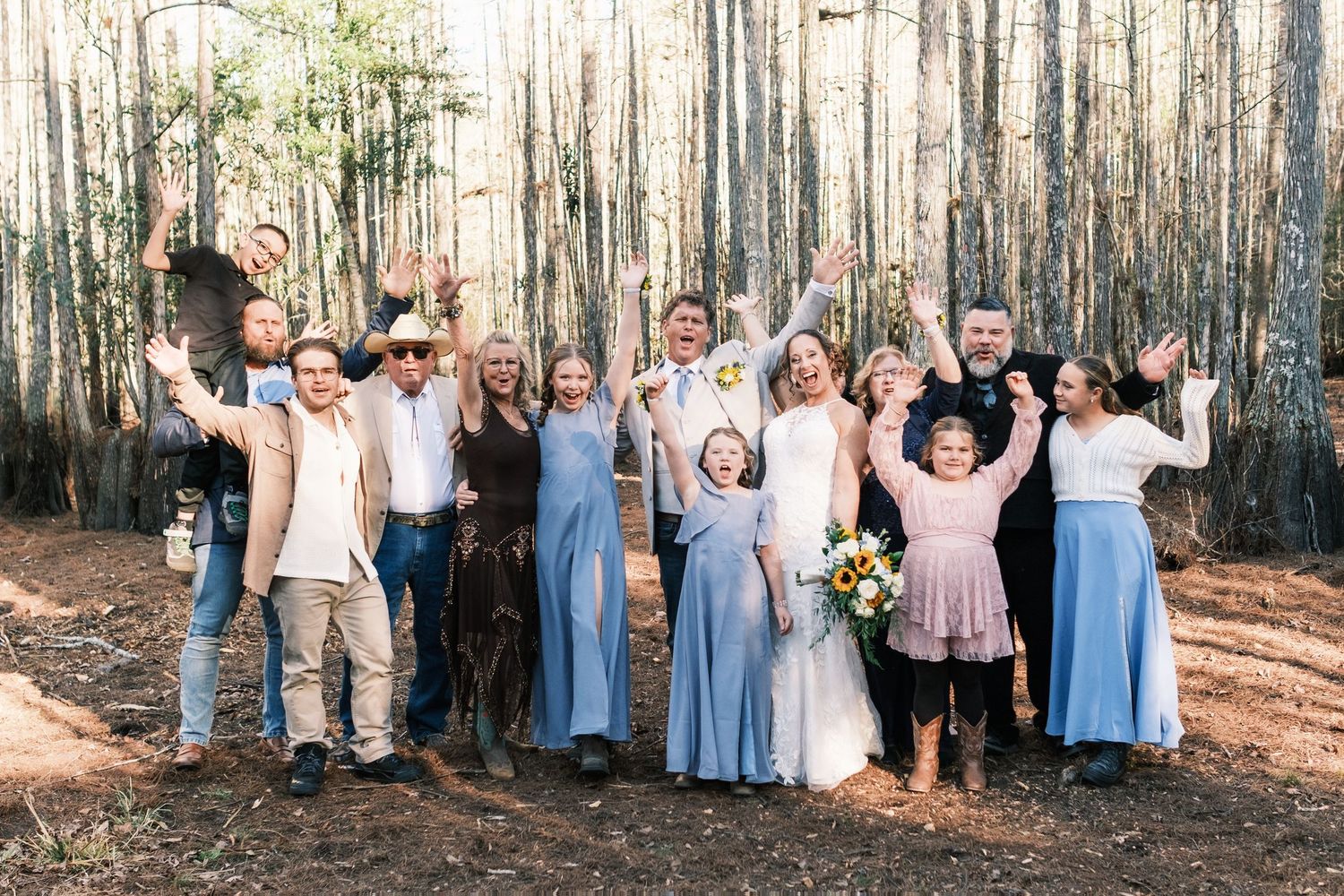 Wedding party celebrating with raised arms in a pine forest, bridesmaids in blue dresses.