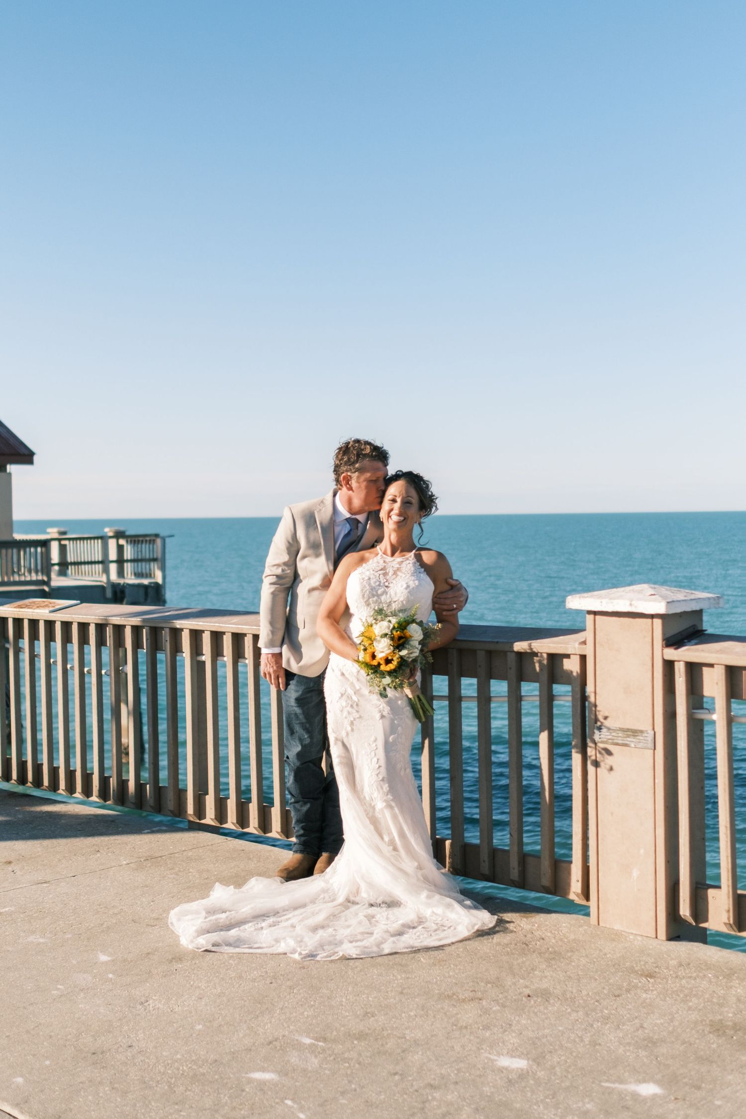 Bride and groom pose romantically on a waterfront pier with ocean views on their wedding day.