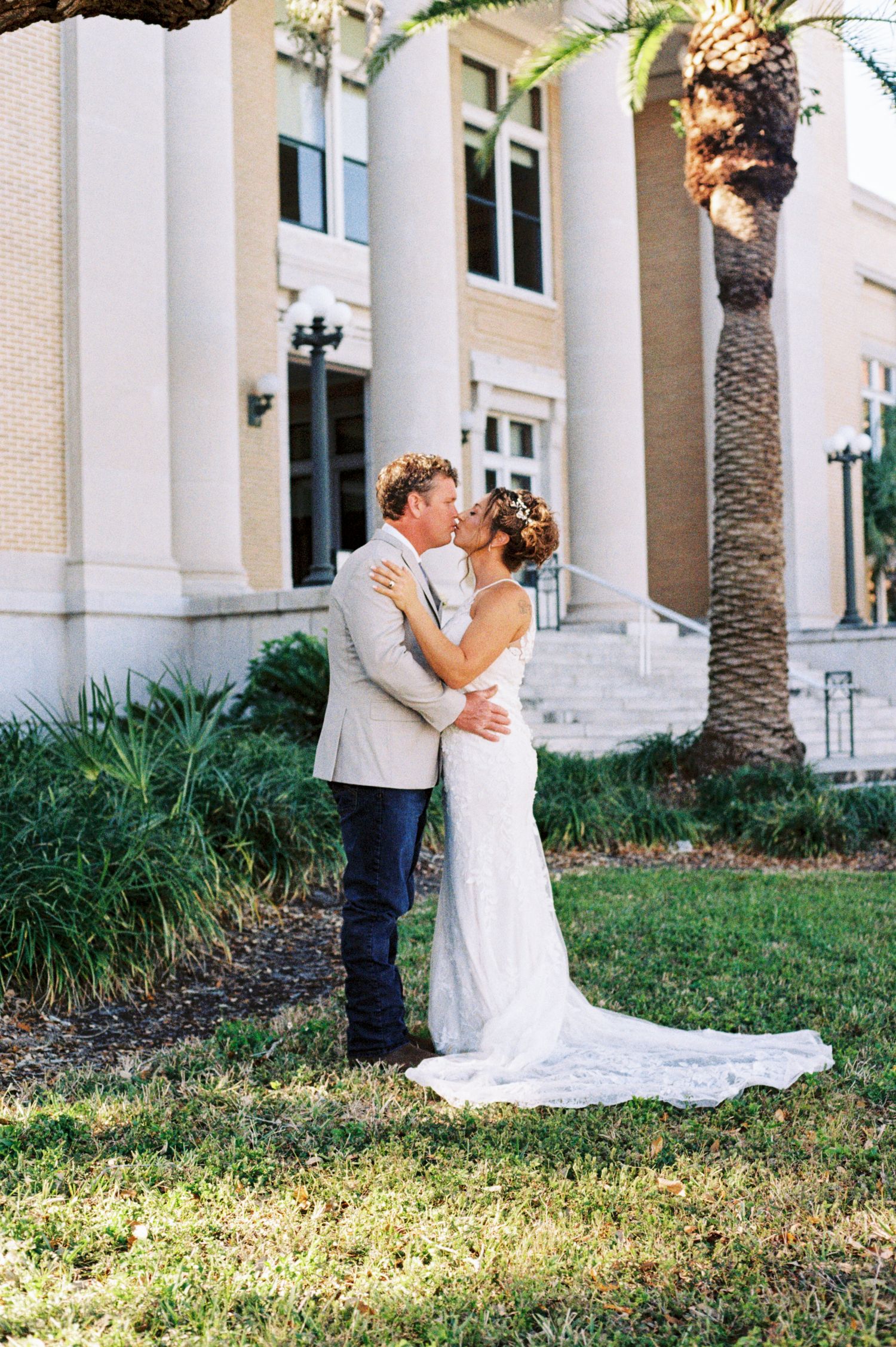 A bride and groom share a passionate kiss on the lawn outside a grand building.
