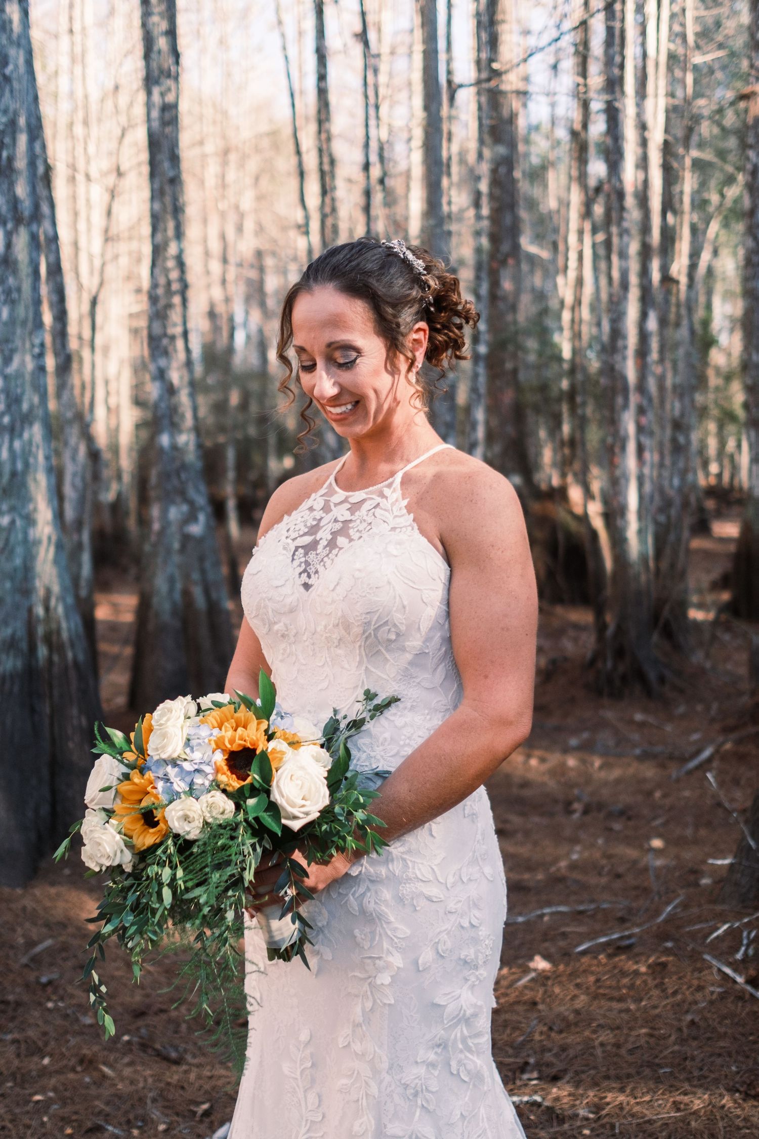 Bride in lace halter wedding gown holding sunflower bouquet smiling in pine forest.