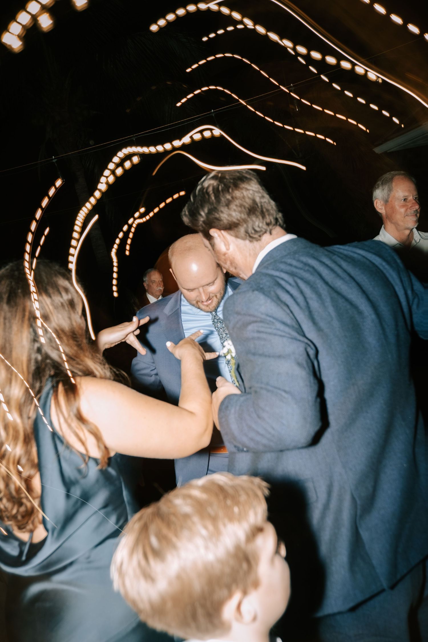 Wedding guests dancing under string lights at an evening reception celebration.