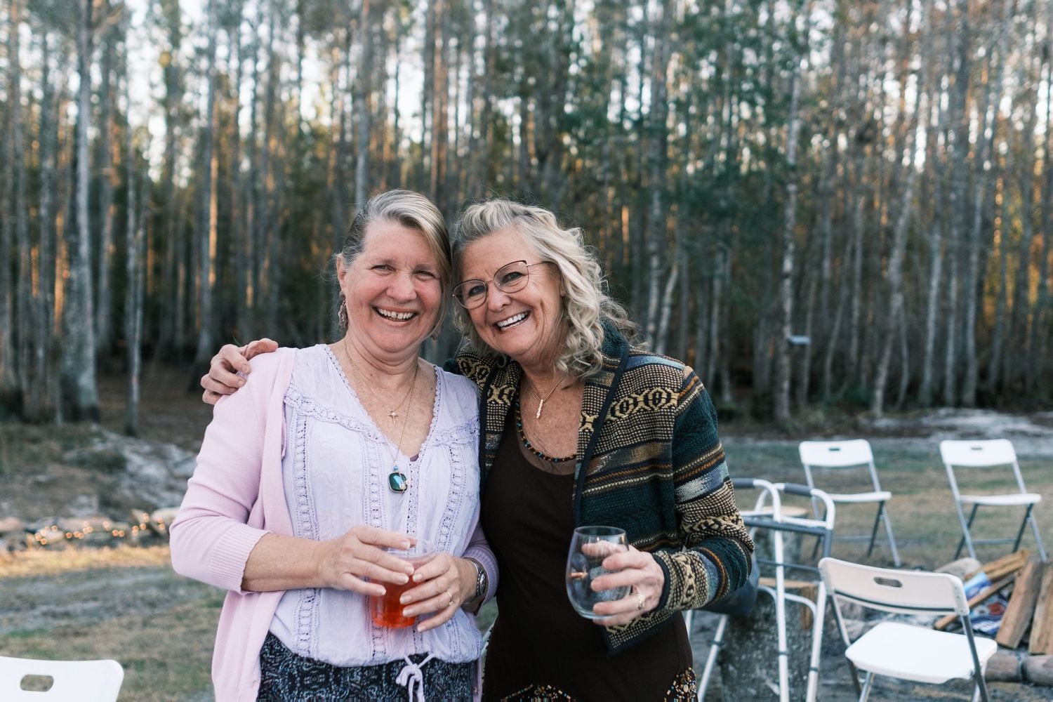 Two smiling women holding drinks embrace outdoors at evening gathering among tall trees.