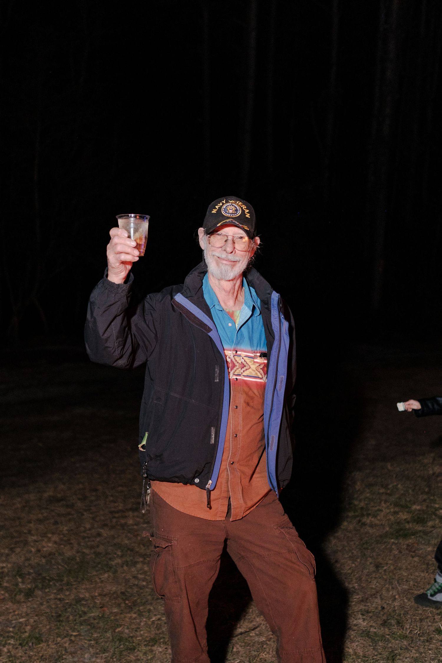 Elderly man in a baseball cap raising a glass in a toast outdoors at night.