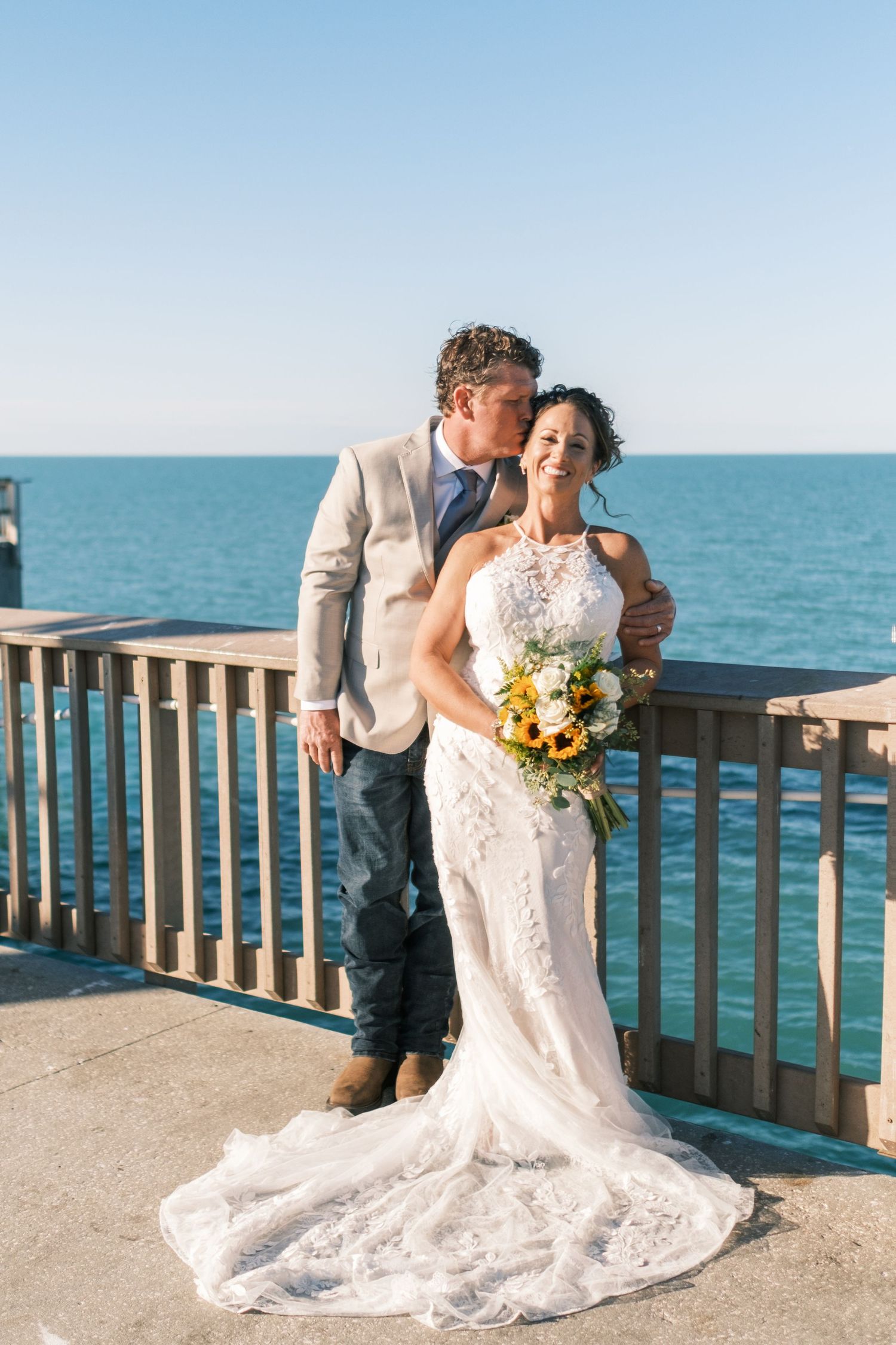 Bride and groom pose on a waterfront pier, groom kissing her cheek as she holds a sunflower bouquet.