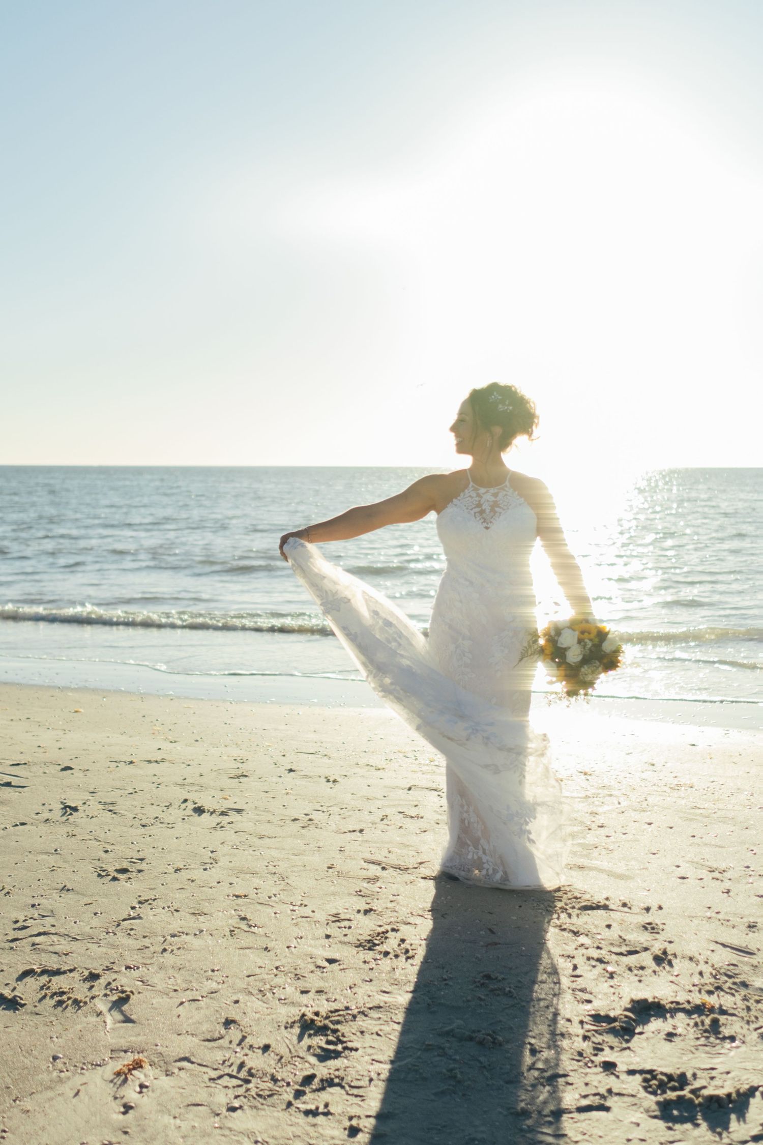 Bride in white lace gown holds bouquet on sunny beach with ocean backdrop at sunset.