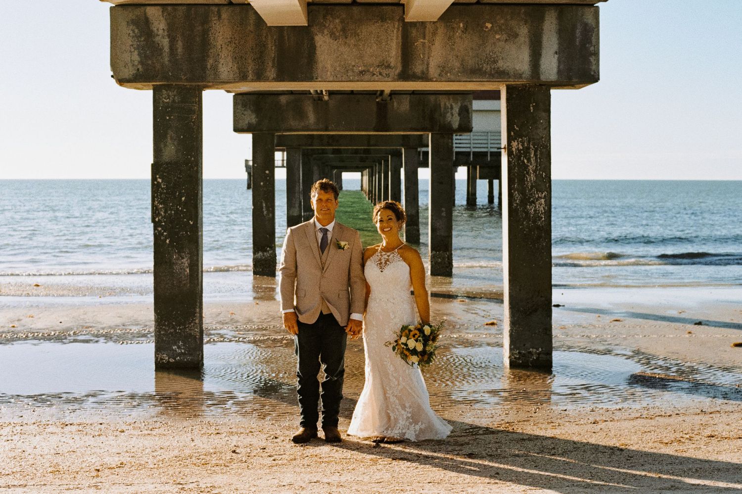 Bride and groom pose under a pier on the beach with ocean in the background.