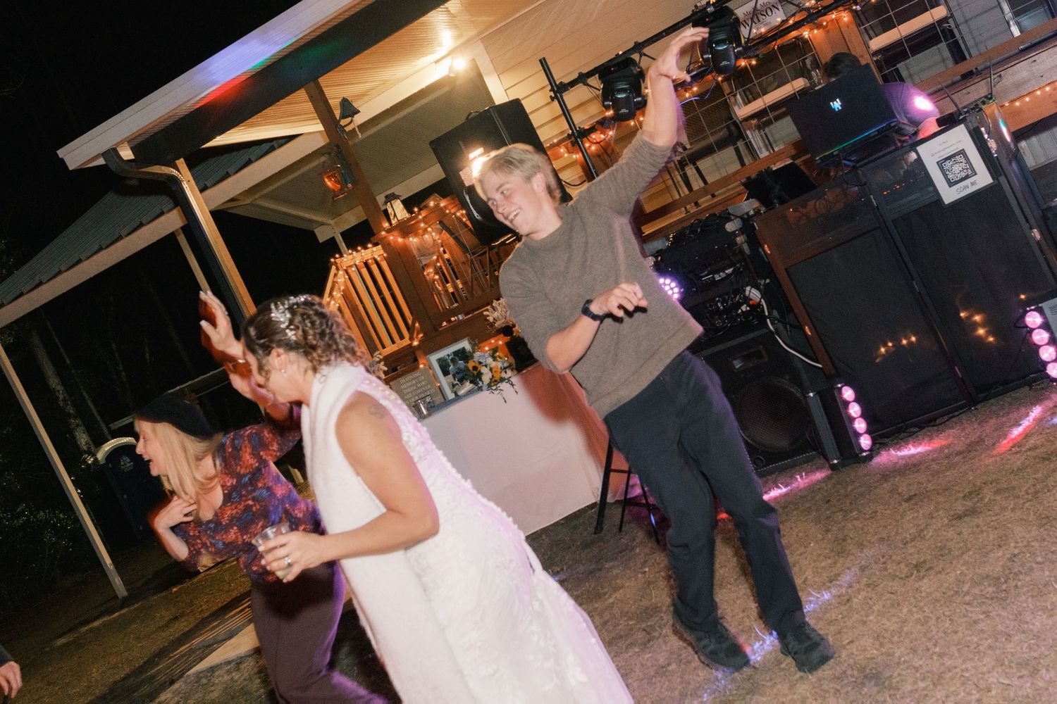 Bride in white gown dances energetically with guests at an outdoor wedding reception at night.