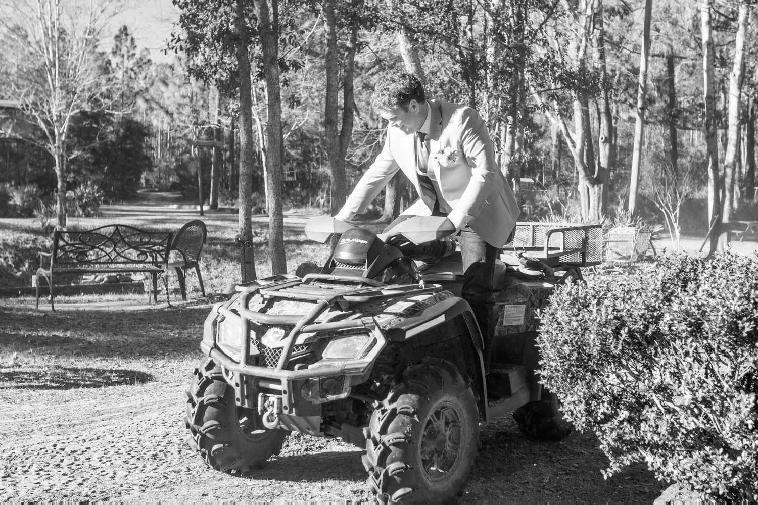 Groom in wedding attire rides an ATV through a wooded outdoor area, black and white photo.