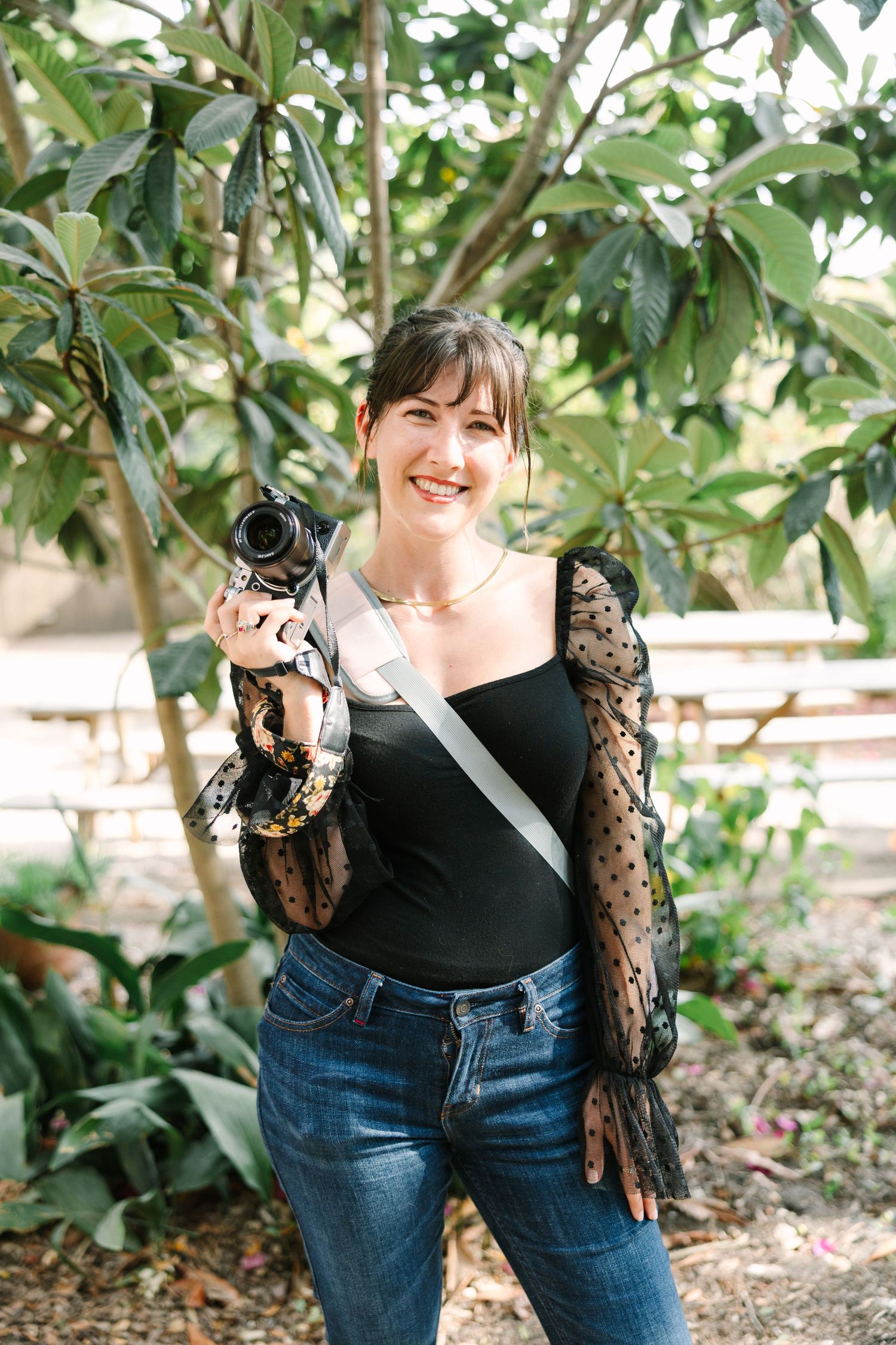 Photographer in a black lace top and jeans holding a camera in an outdoor garden setting.