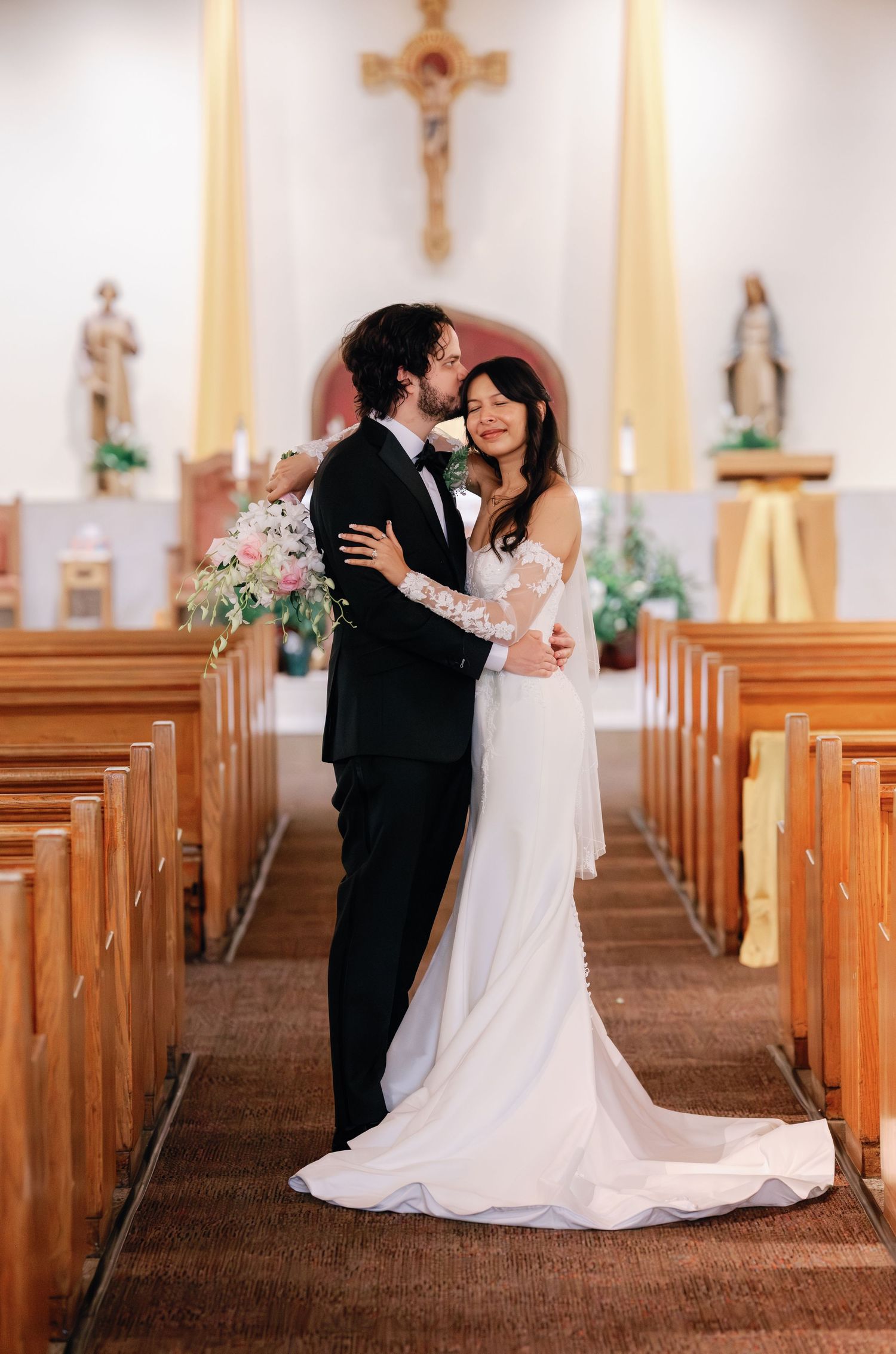 A bride and groom share a romantic kiss in a church aisle, she in a white gown holding flowers, he in a black tuxedo.