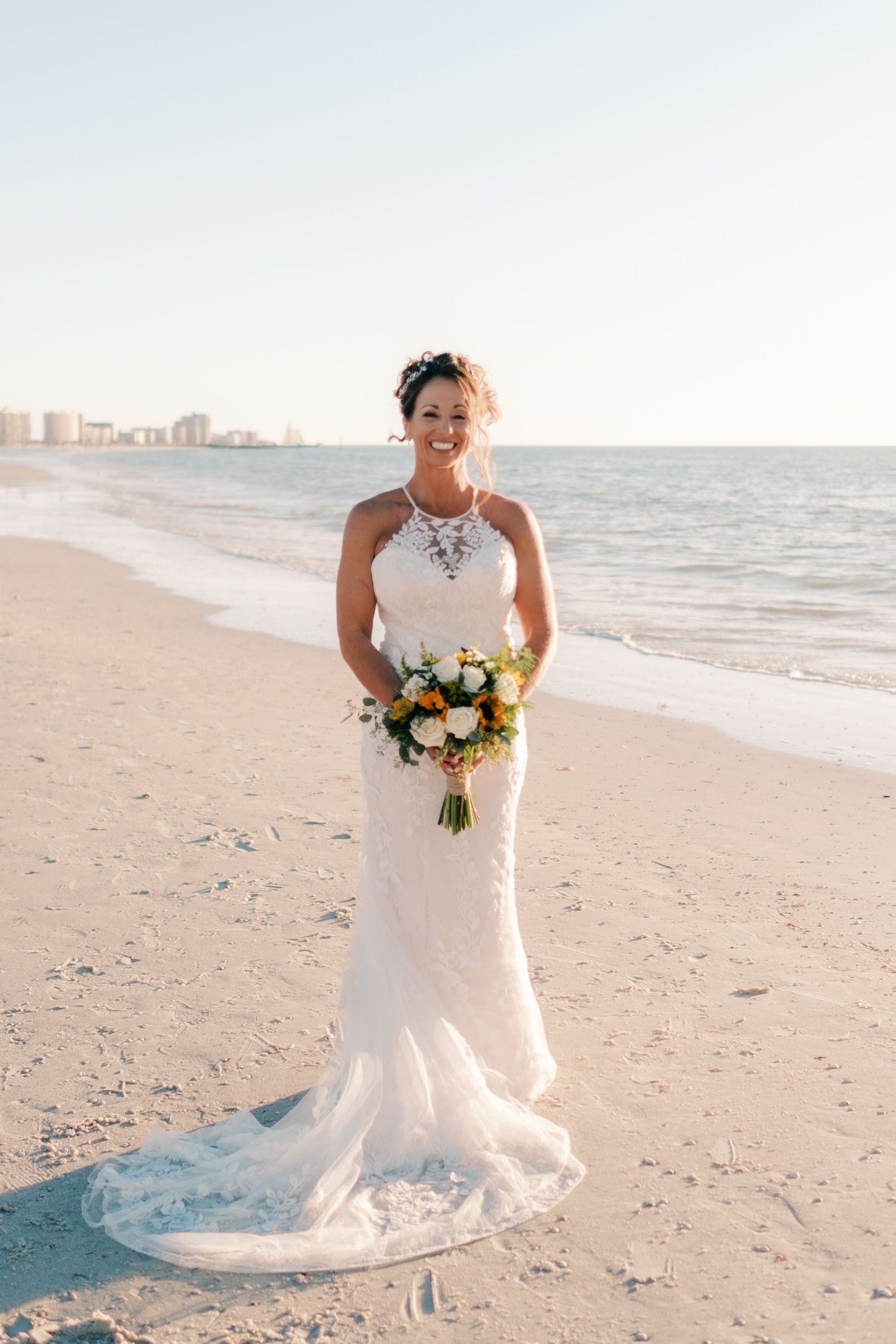 Radiant bride in lace halter gown holds sunflower bouquet standing on sunny beach shoreline.