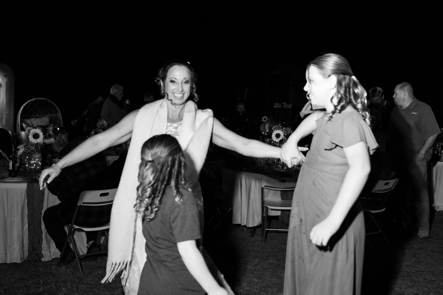 Black and white photo of bride dancing joyfully with two young girls at a wedding reception.