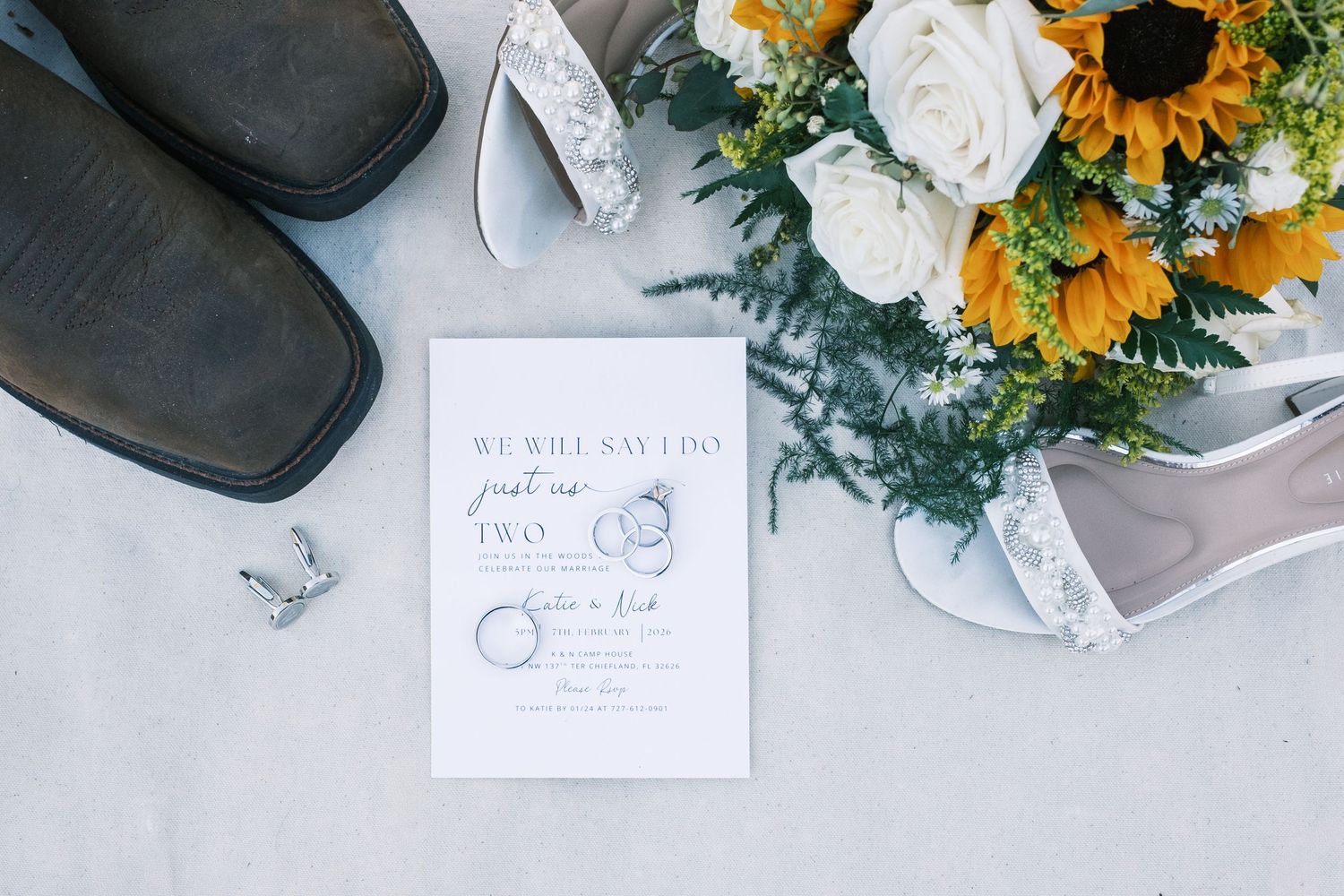 Wedding flat lay featuring sunflower bouquet, cowboy boots, heels, rings, and elegant save the date card.