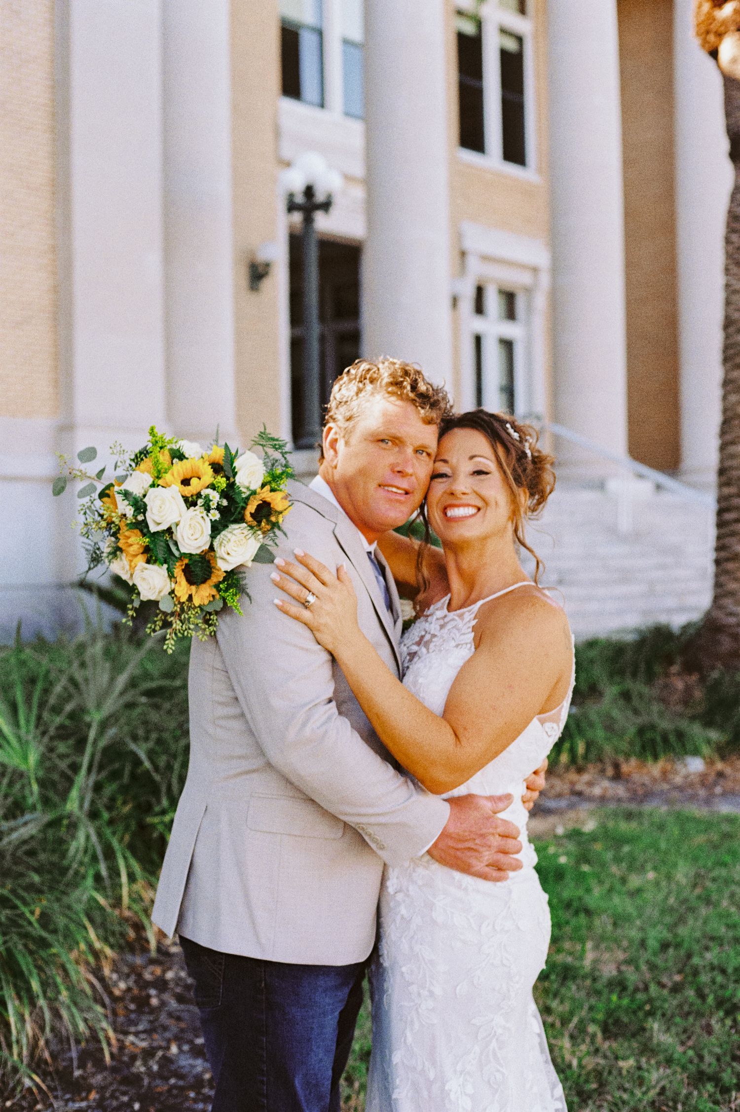 Smiling bride and groom embrace outside classical building holding sunflower and white rose bouquet.