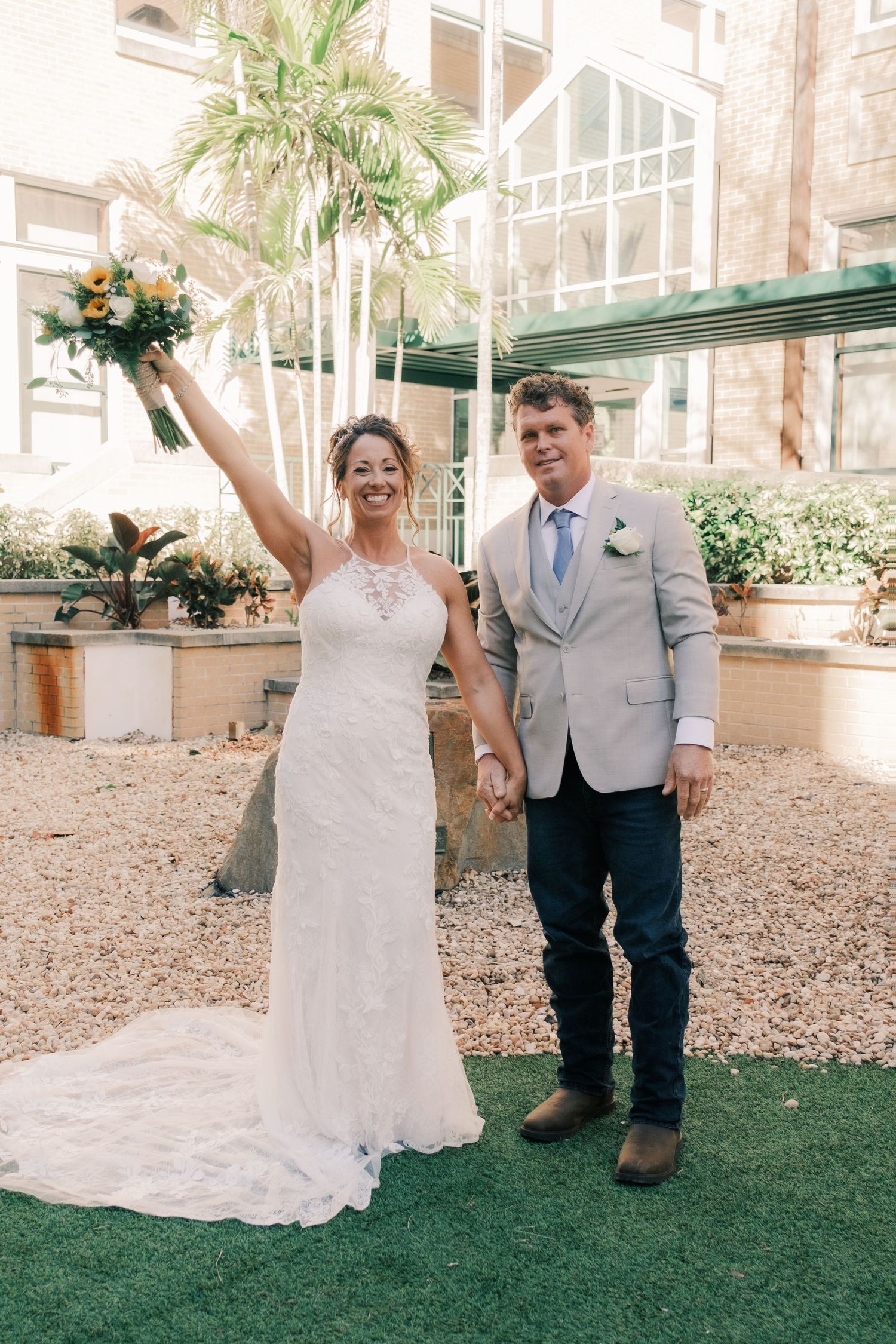 Joyful bride raising bouquet with groom in courtyard with palm trees.