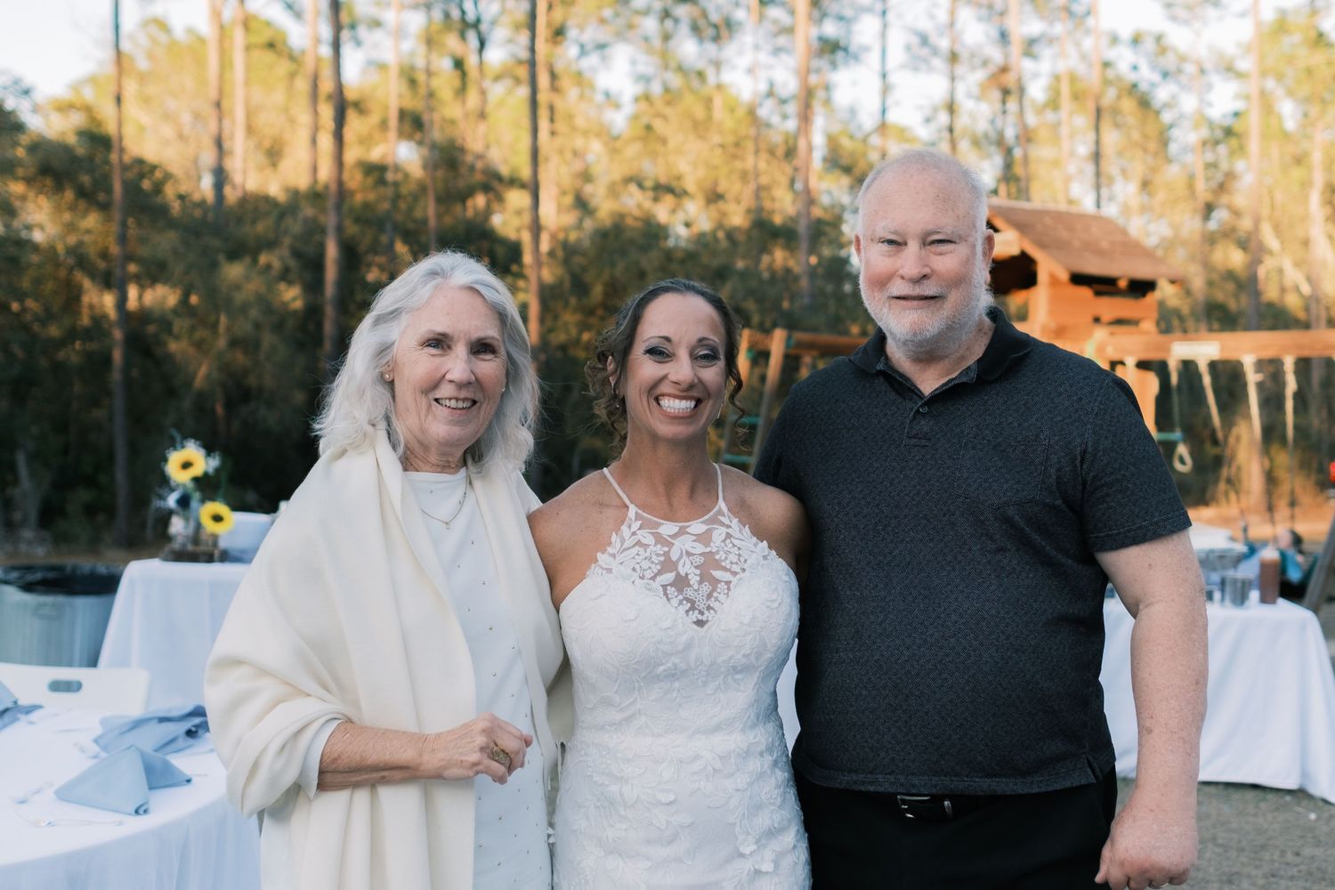 Bride in halter lace gown poses with older couple at outdoor wedding reception near sunflower decorations.