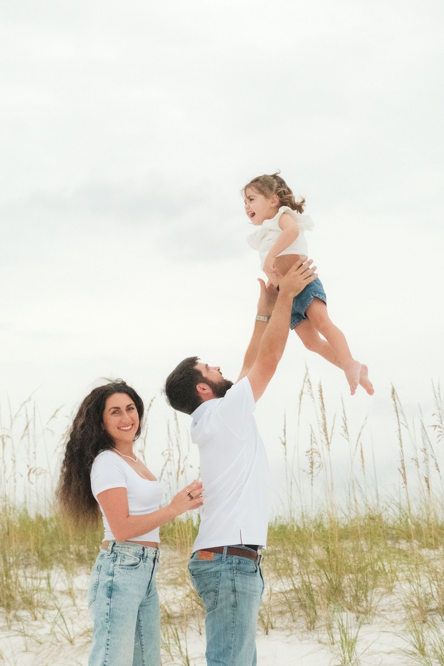A couple in casual denim and white outfits playfully interacts on a sandy beach with sea grass in the background.