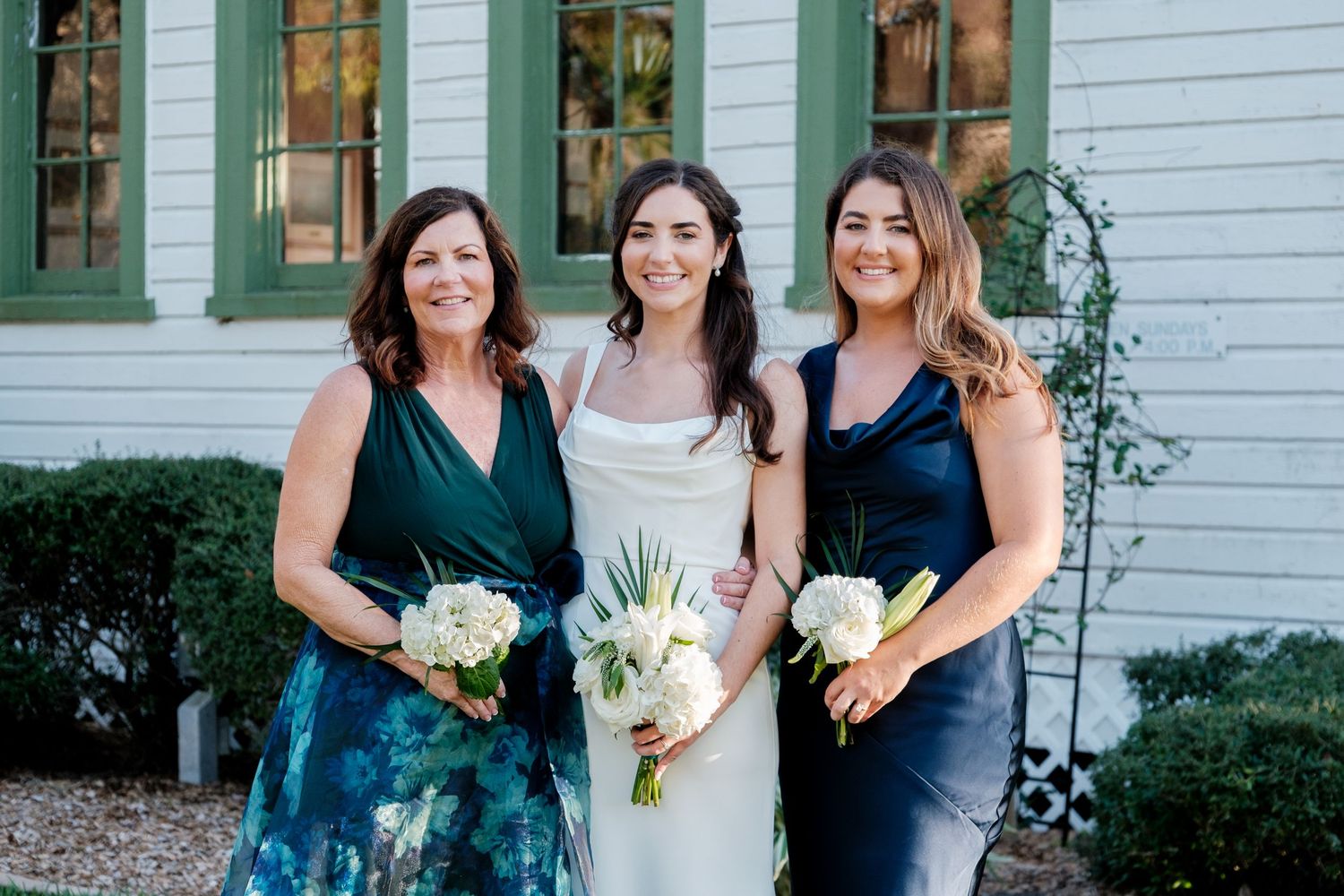 Three women pose together outside a white house with green trim, holding wedding bouquets.