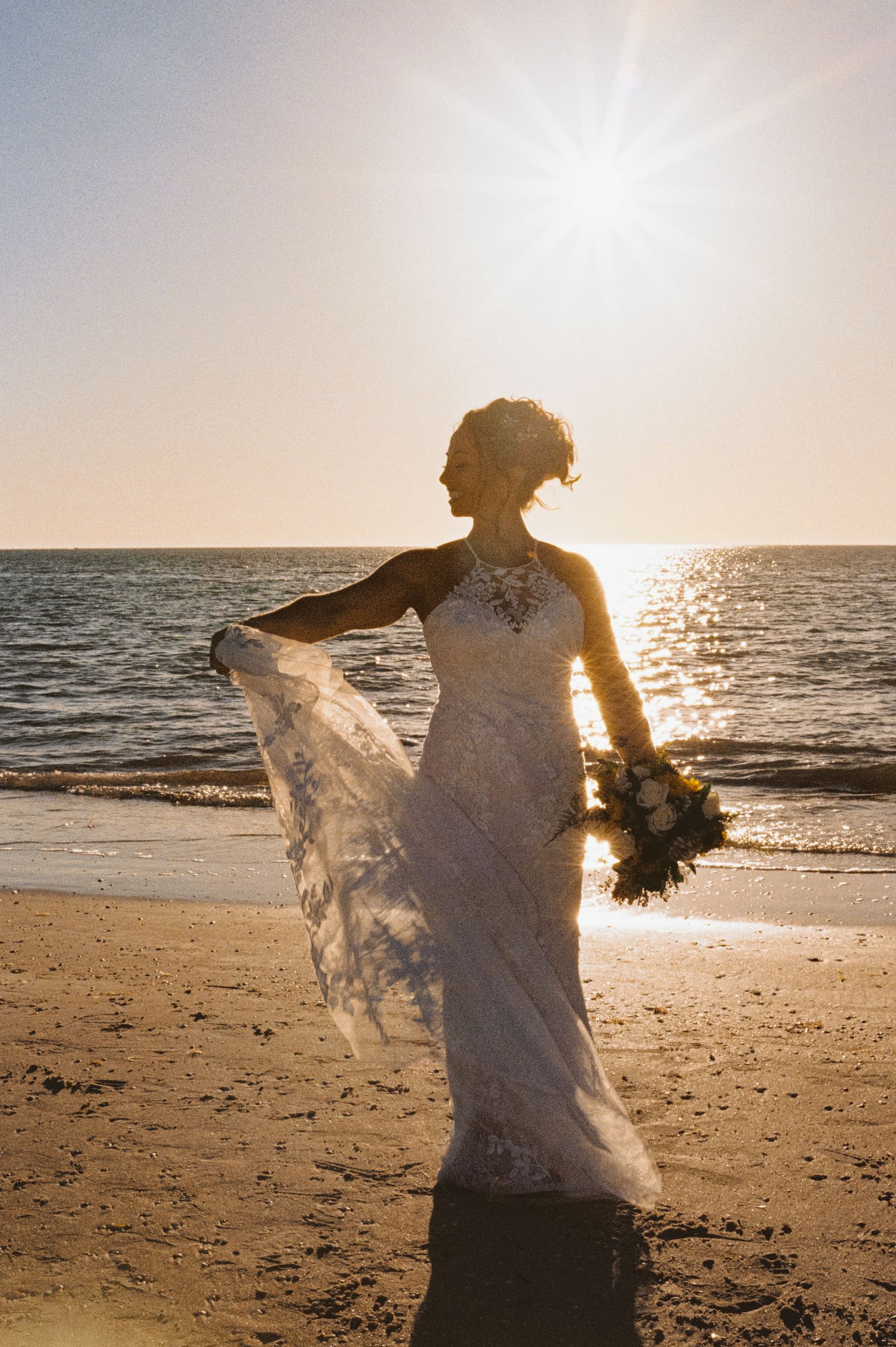Bride in lace gown holds bouquet while her veil flows in the breeze on a sunlit beach at golden hour.