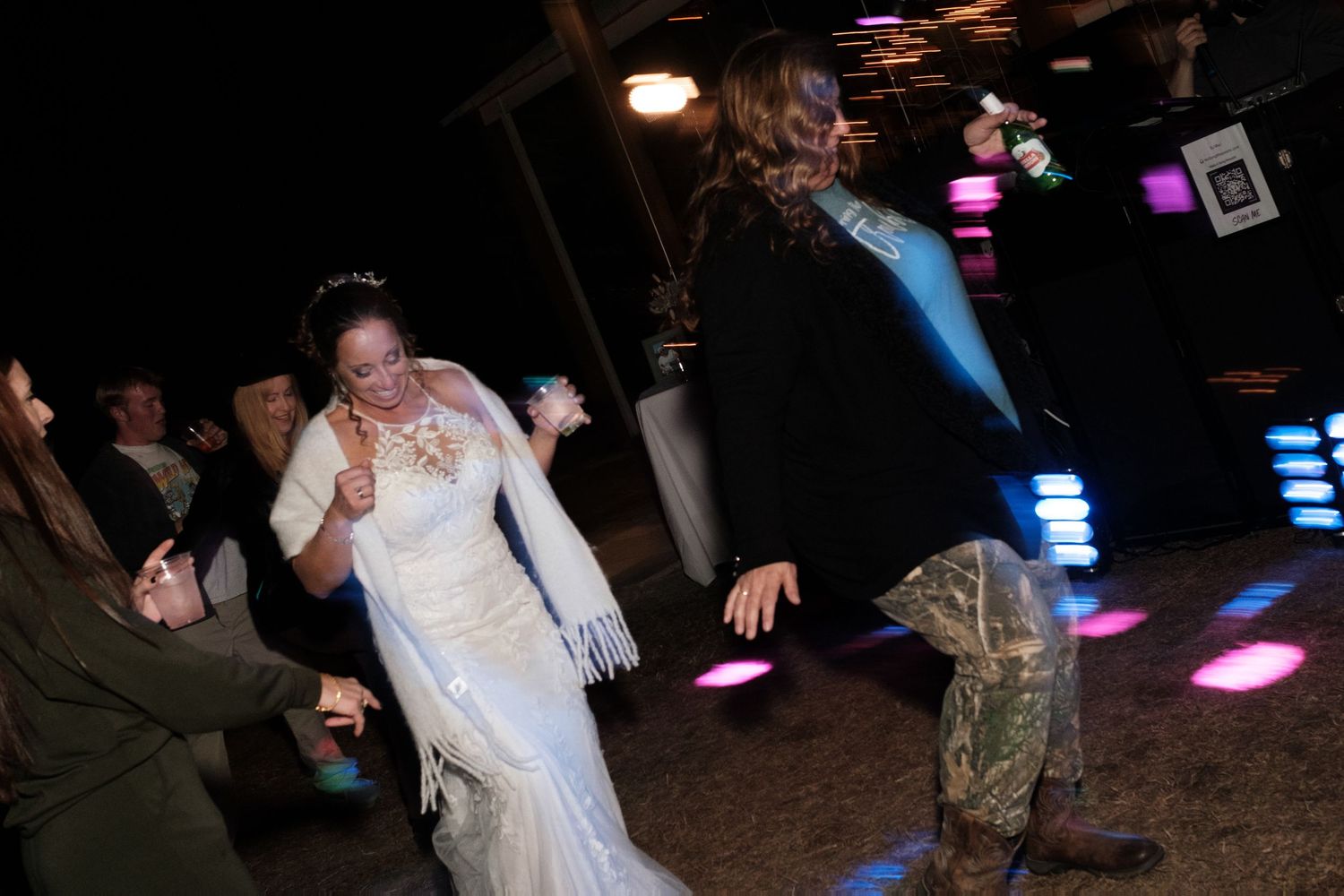 Bride in white gown with fur wrap dances energetically at lively nighttime wedding reception.