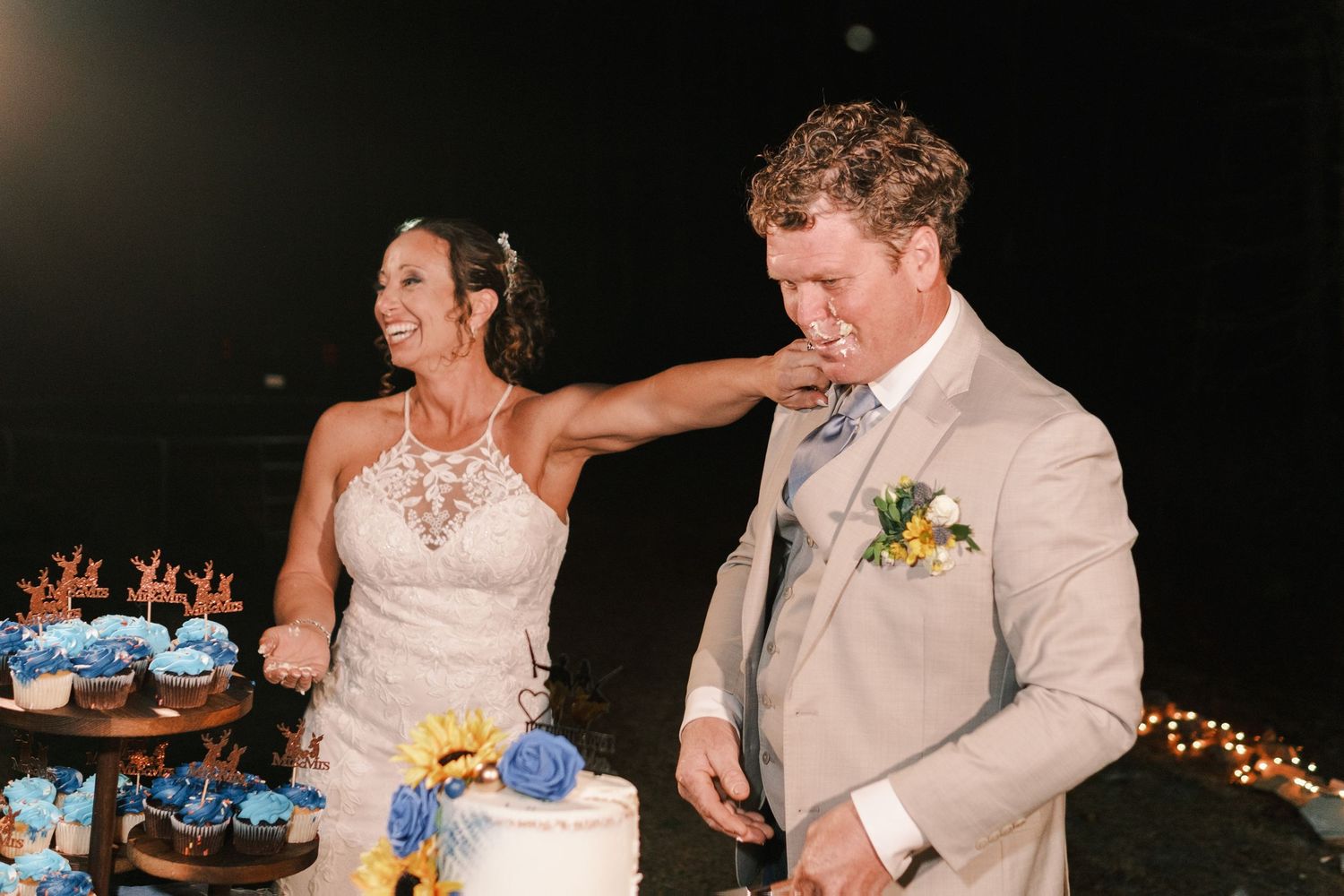 Bride smiling as groom tastes wedding cake during outdoor evening reception.