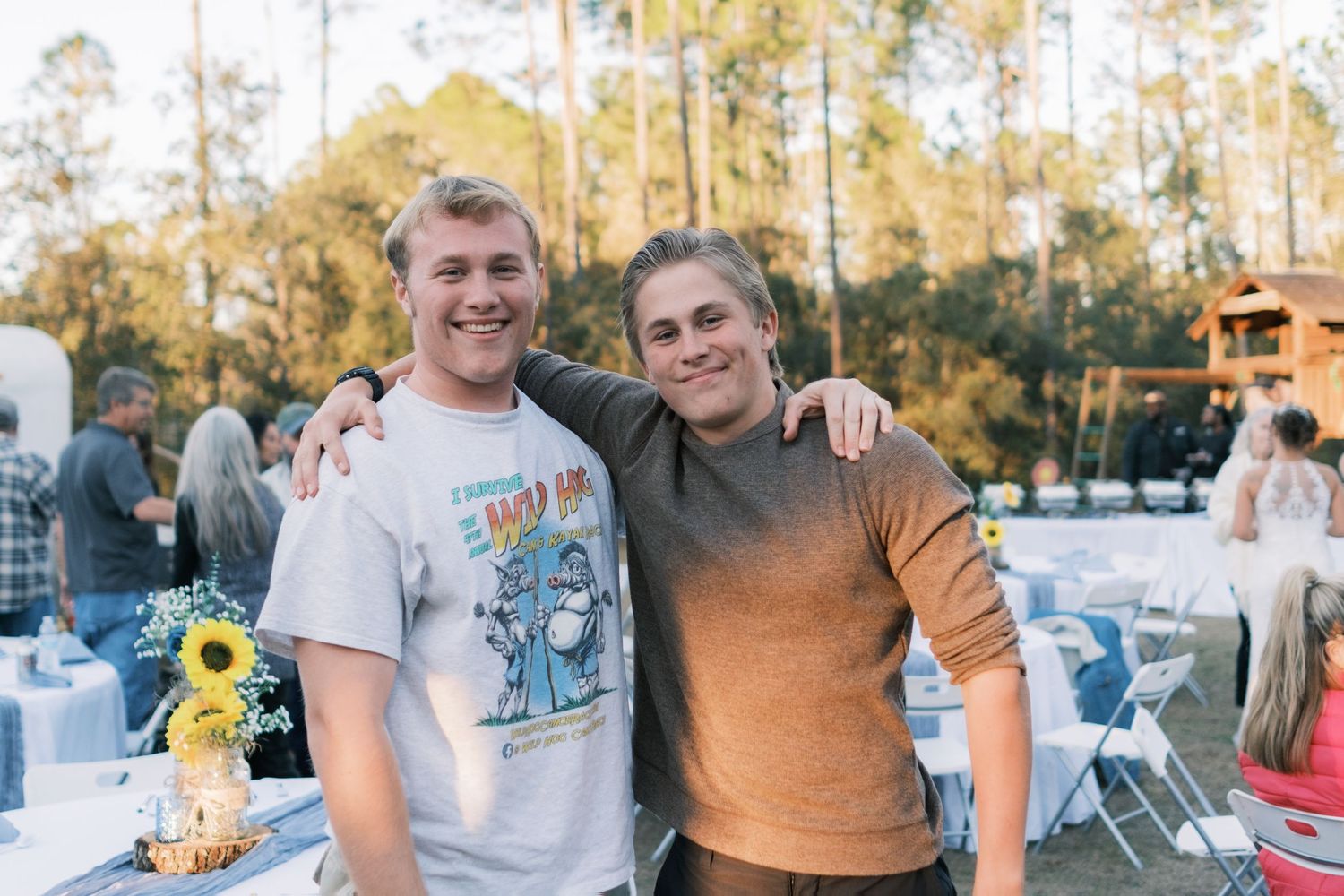 Two young men smile with arms around each other at an outdoor wedding reception.