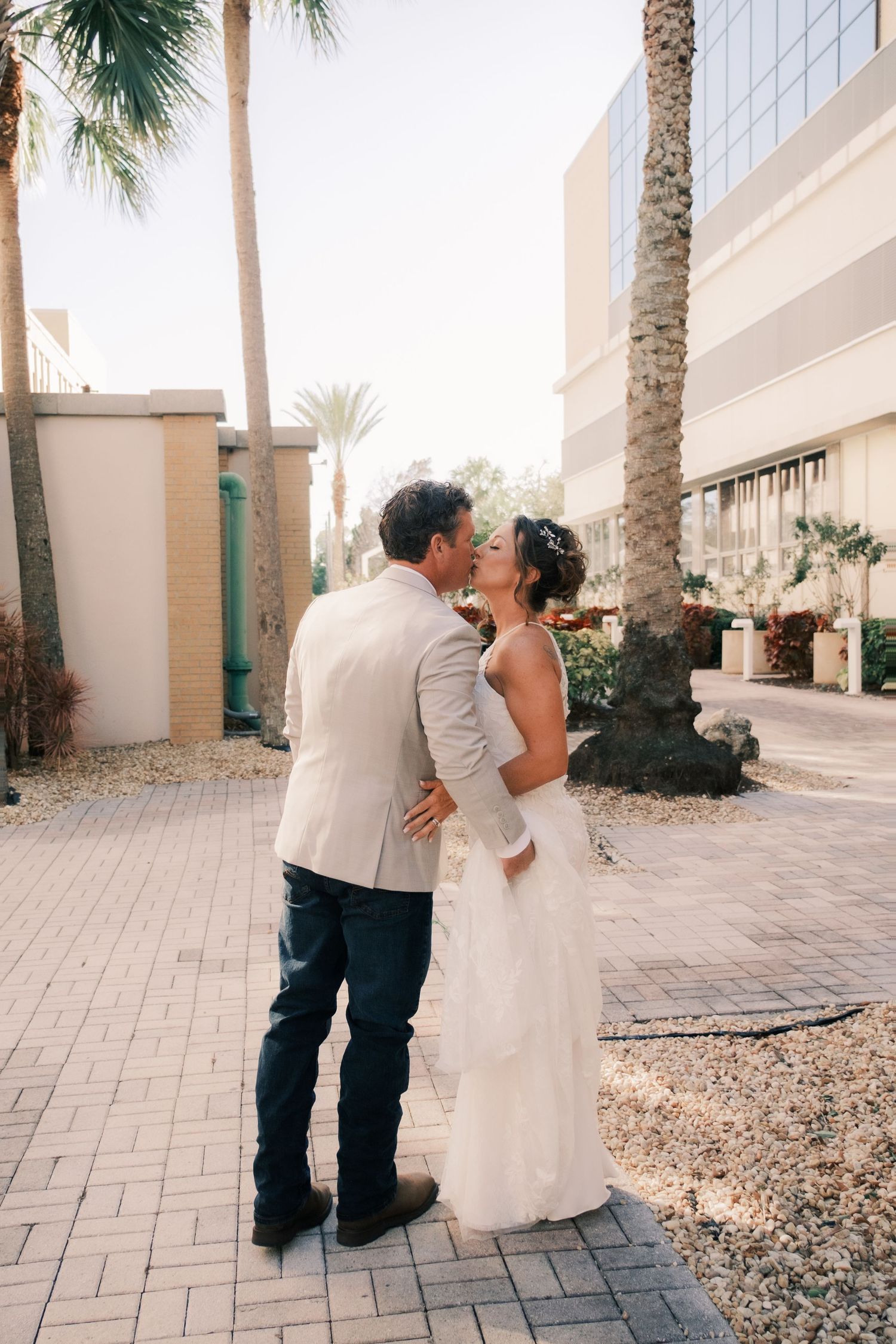 Bride and groom share a romantic kiss on a sunny paved walkway surrounded by palm trees.
