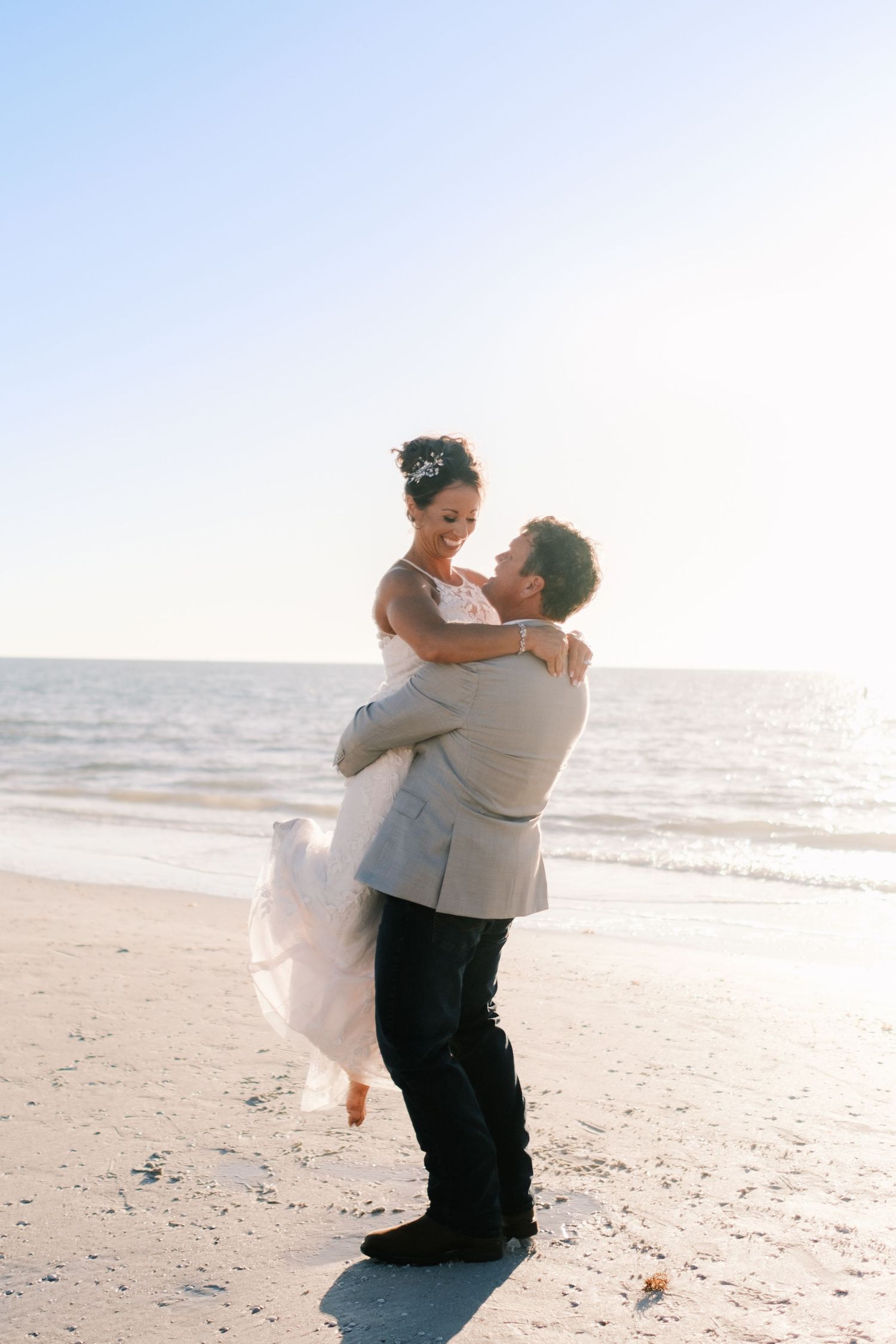 Groom lifting bride in white dress on a sunny beach with ocean waves in the background.