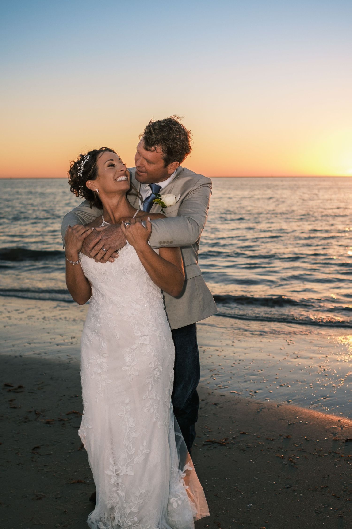 Bride and groom share romantic sunset beach embrace, groom kissing bride's cheek in wedding attire.