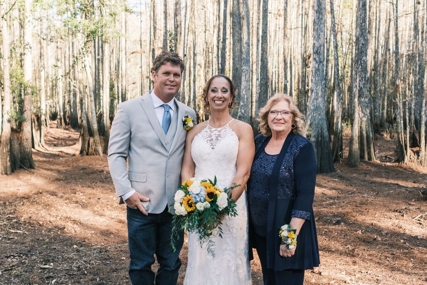 Bride in lace gown poses with groom in gray suit and older woman in navy dress in a wooded forest setting.
