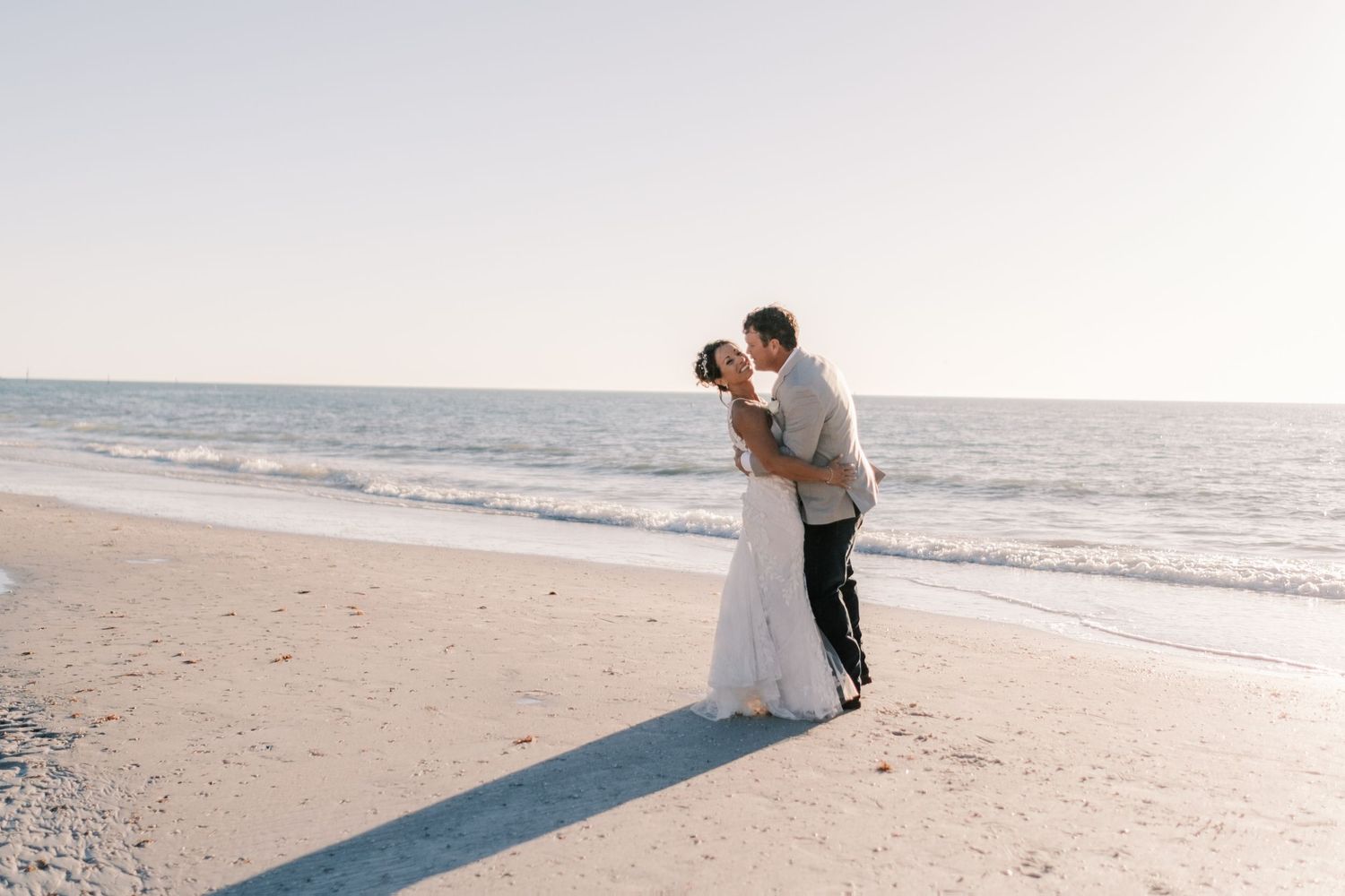 Bride and groom share a kiss on a sandy beach at sunset during wedding.