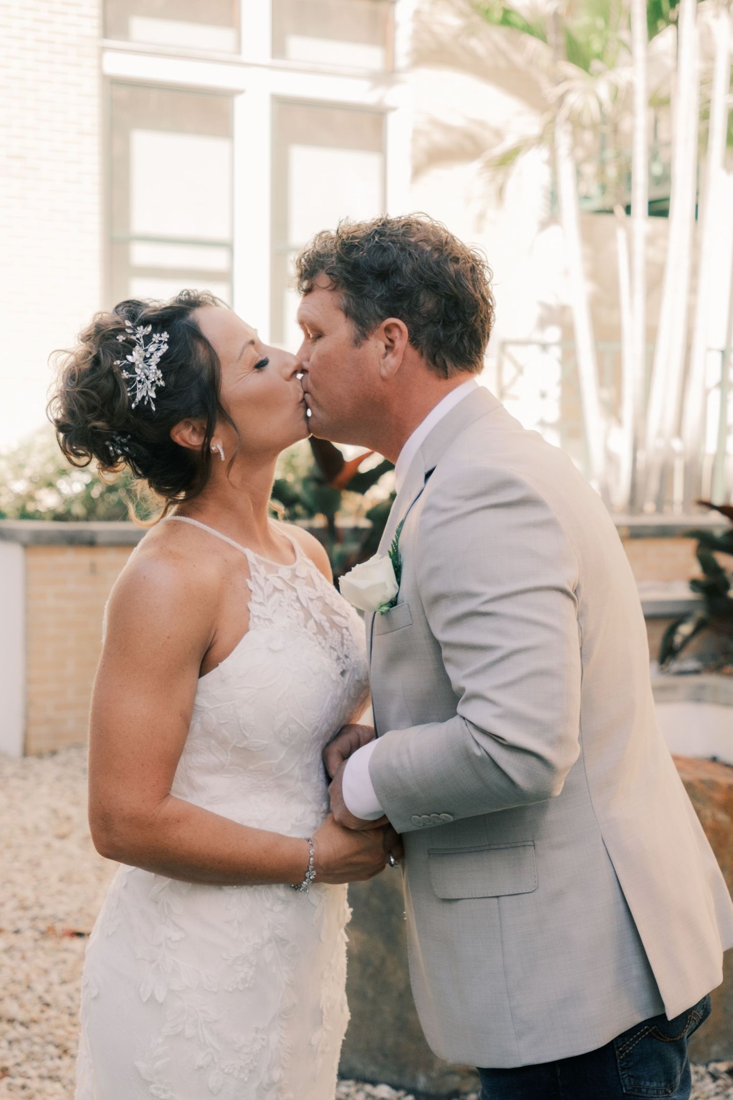 Bride and groom share a romantic first kiss outdoors, bride wearing lace dress with floral hair accessory.
