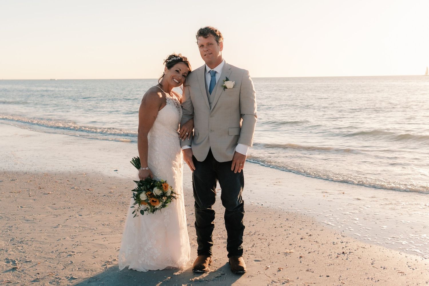 Bride and groom smile together on the beach at sunset with ocean waves behind them.