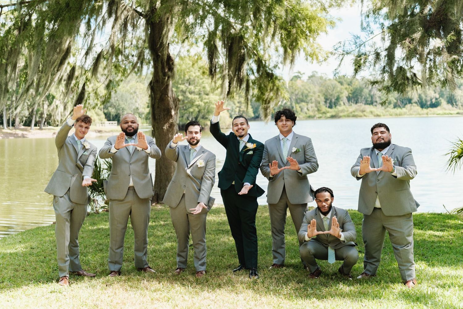 Groomsmen in gray suits posing playfully by a lakeside with Spanish moss trees in the background.
