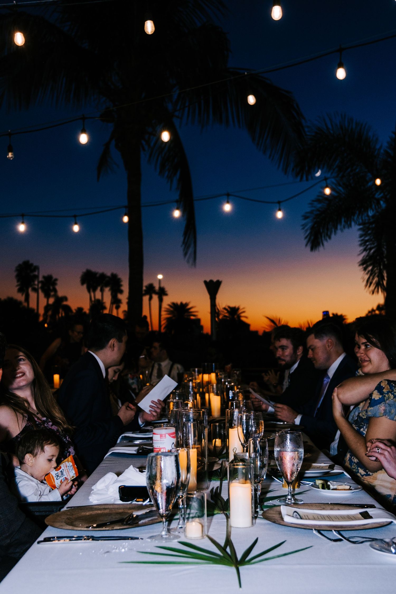 Guests seated at an outdoor dinner table under string lights at dusk with palm trees silhouetted against an orange sunset sky.