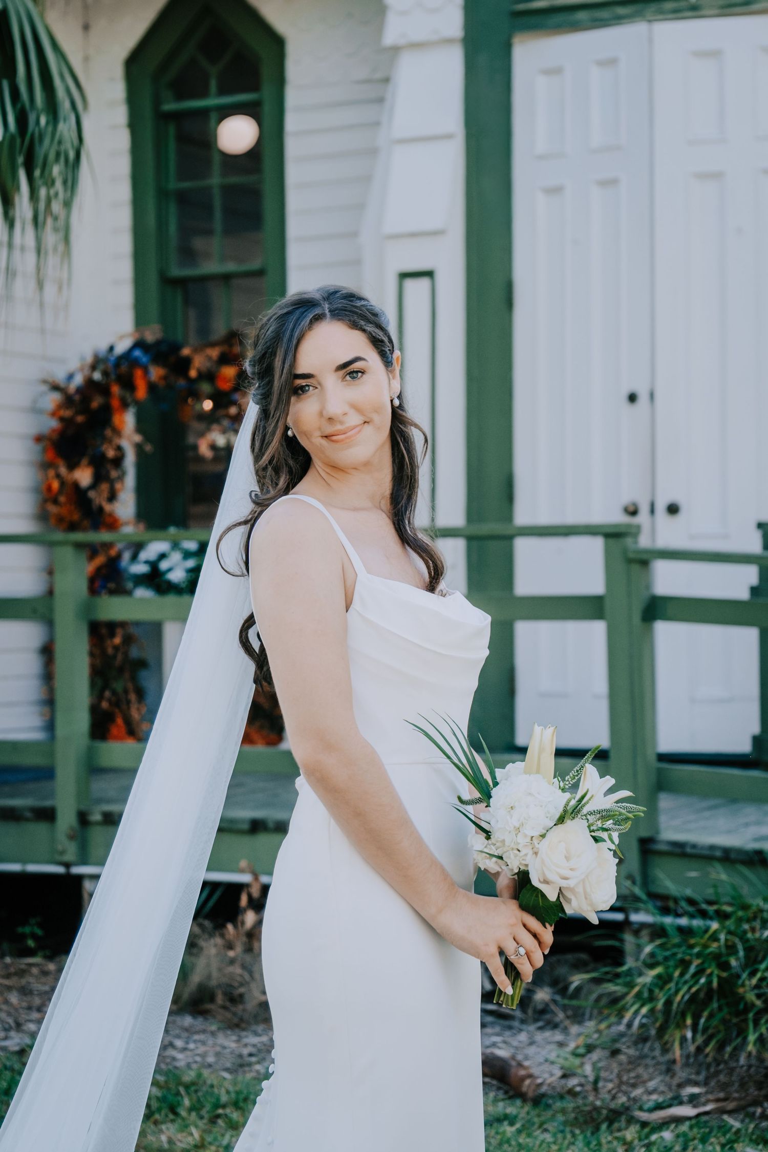 A bride in an elegant white dress holds a bouquet outside a historic white building with green accents.