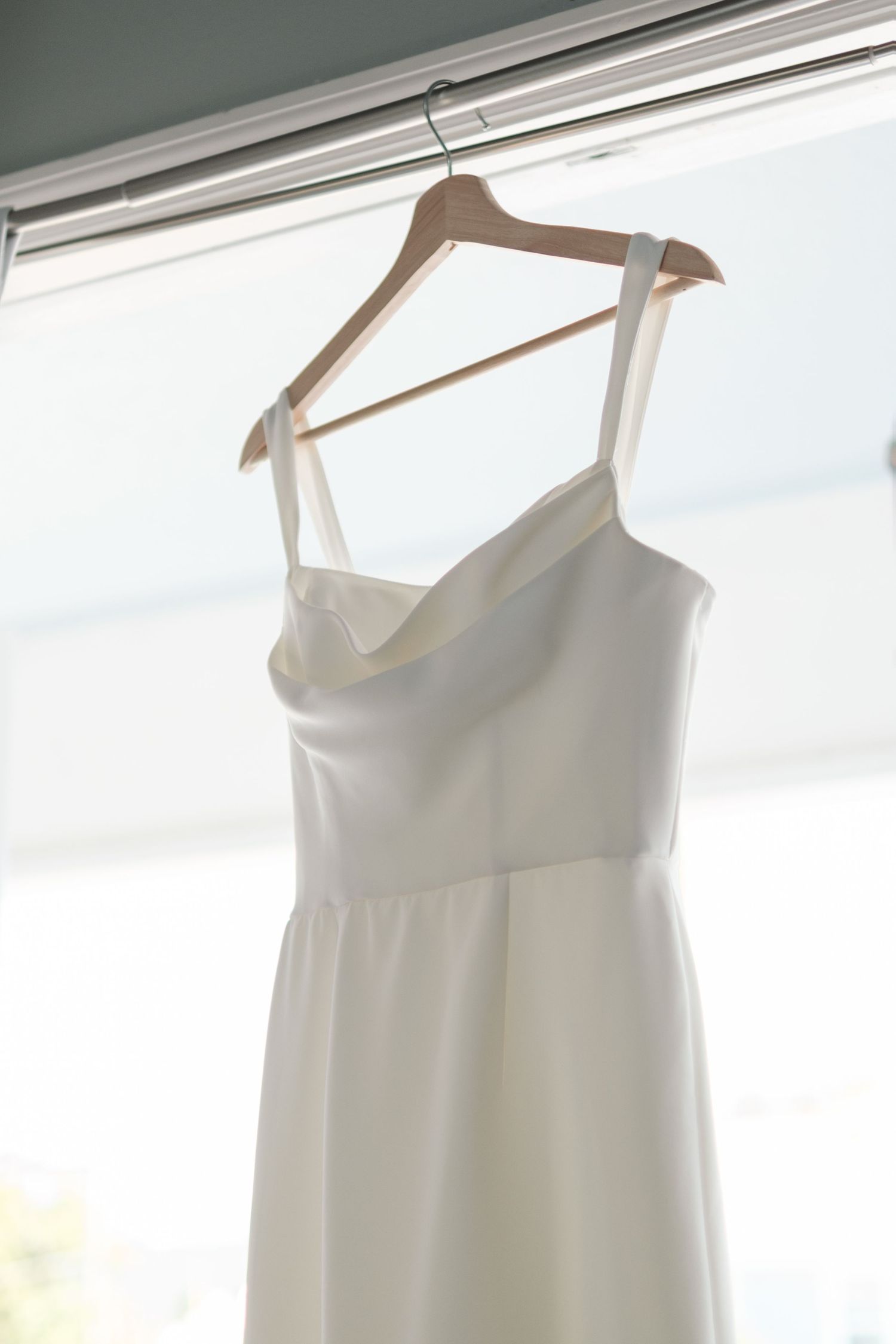 An elegant white wedding dress hangs on a wooden hanger against a bright window.