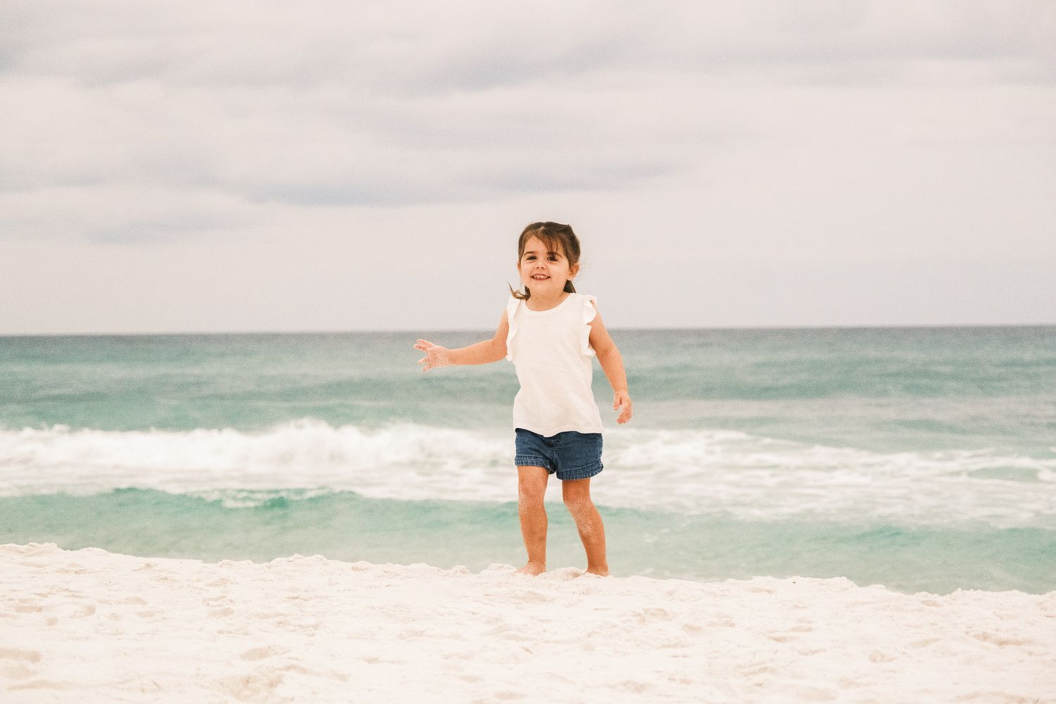 Young child in a white dress plays joyfully at the edge of the ocean waves on a sandy beach.