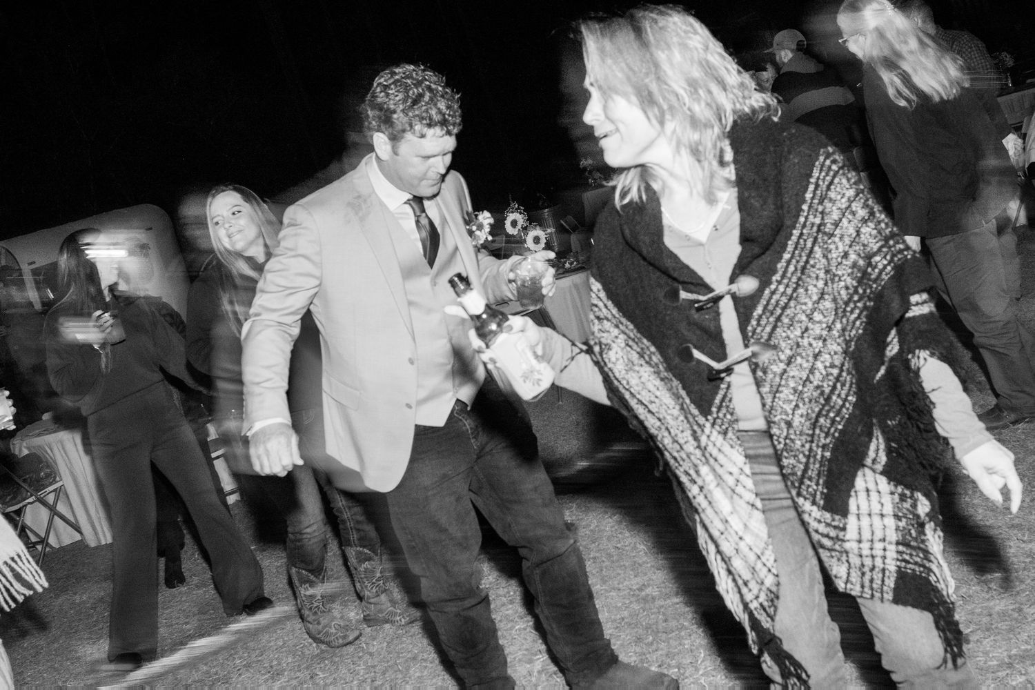 Black and white photo of wedding guests dancing and celebrating outdoors at a nighttime reception.