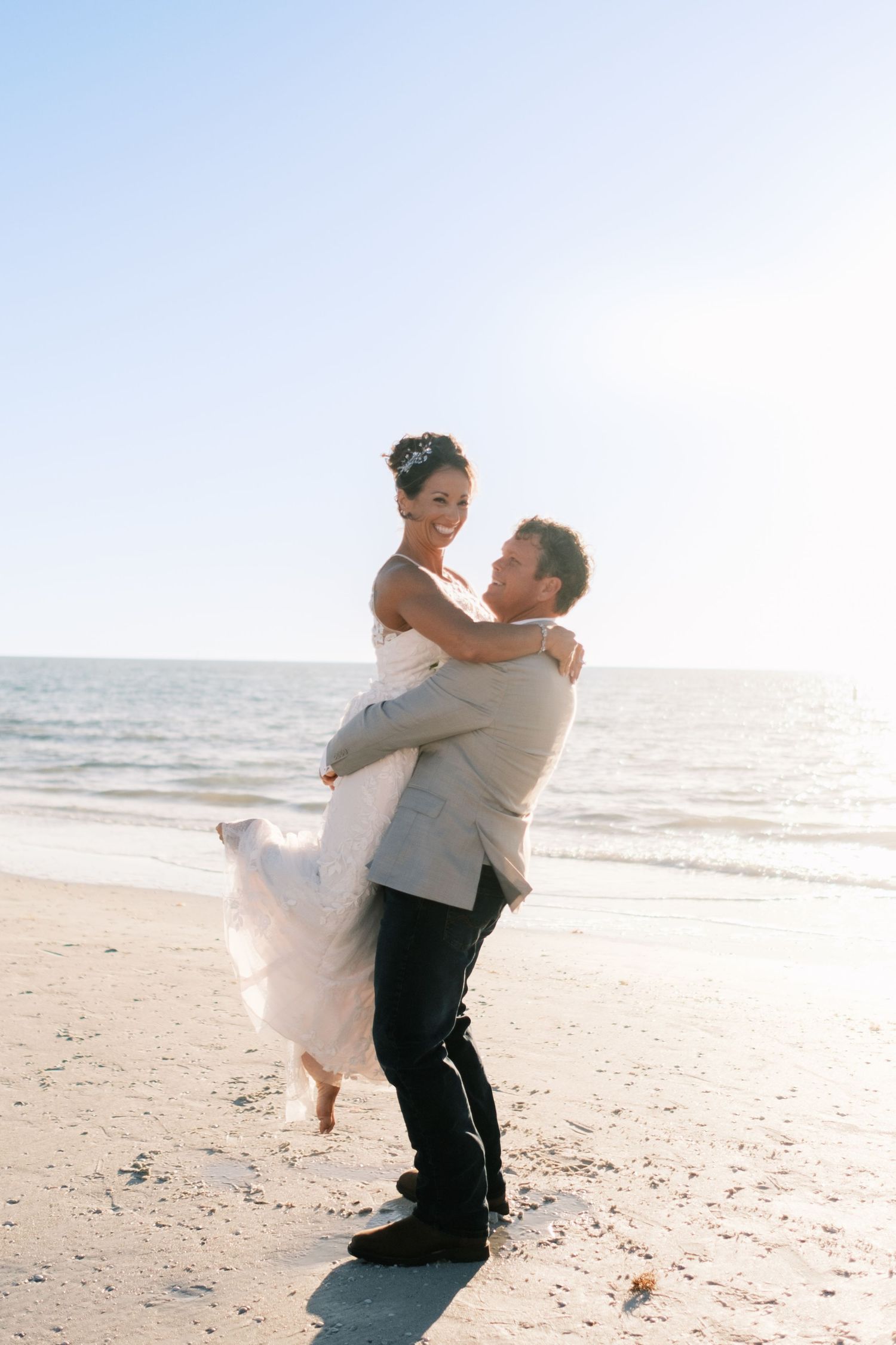 Groom lifts smiling bride in white gown on sunny beach with ocean backdrop.
