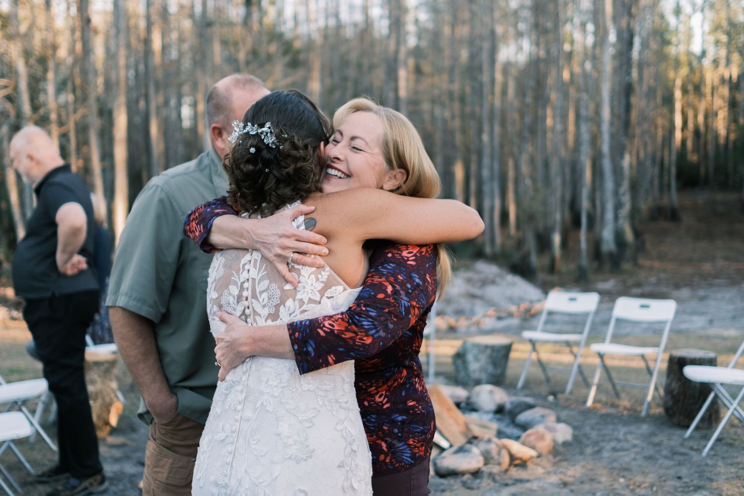 Bride in lace gown receives warm hug from blonde guest at outdoor woodland wedding reception.