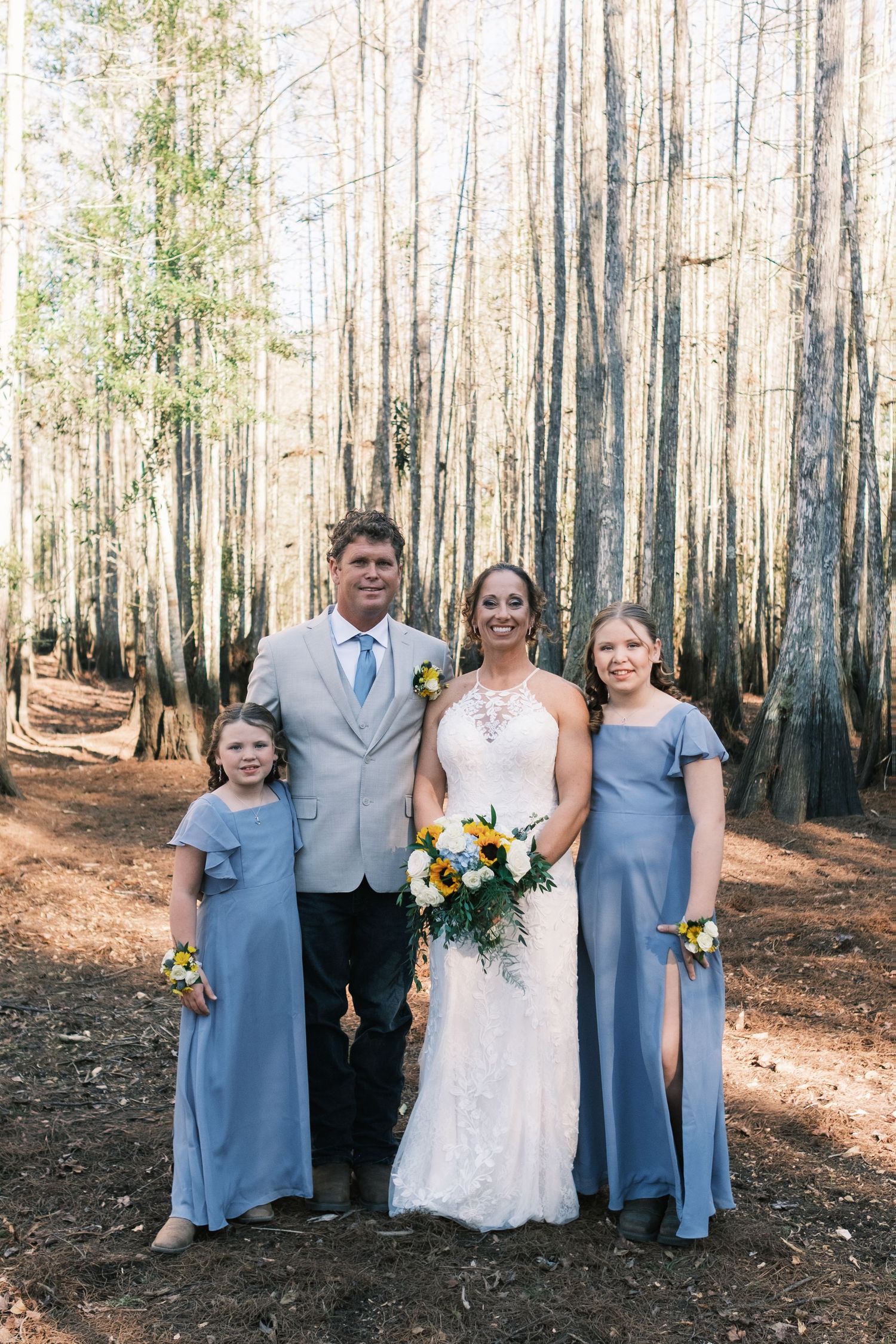 Bride with bouquet poses with groom and two girls in blue dresses in a sunlit pine forest.