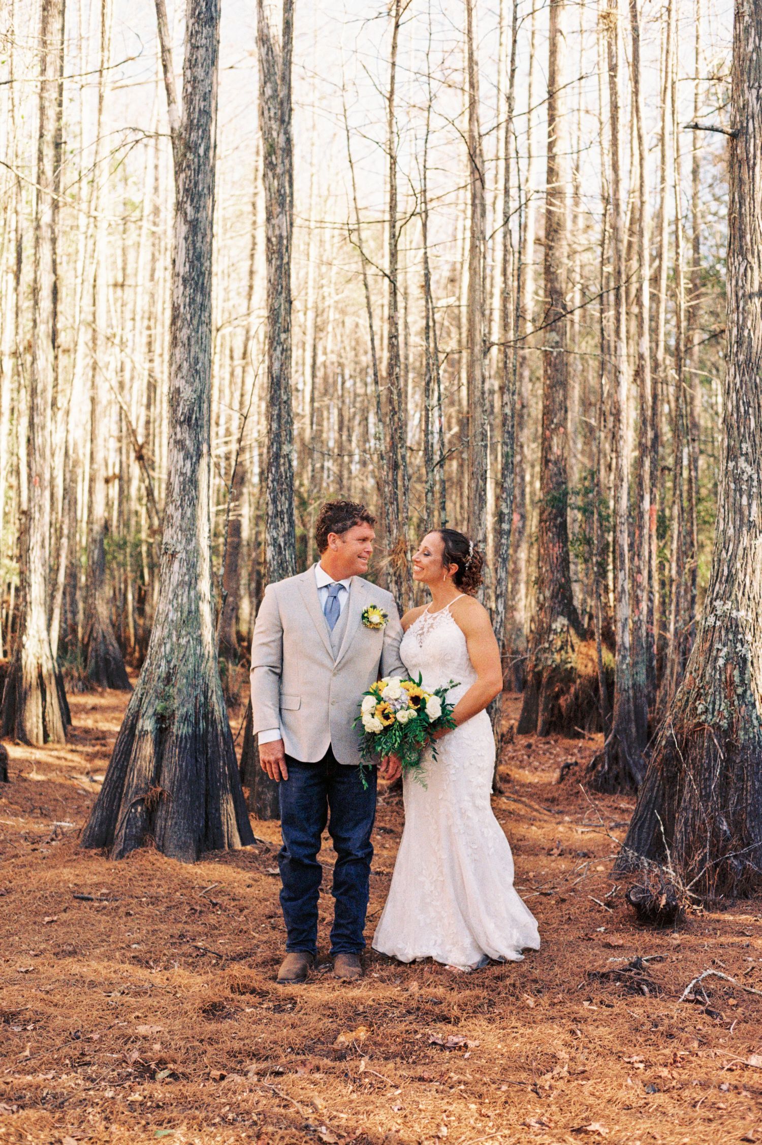 Bride in lace gown and groom in tan blazer share a loving gaze in a sunlit forest setting.