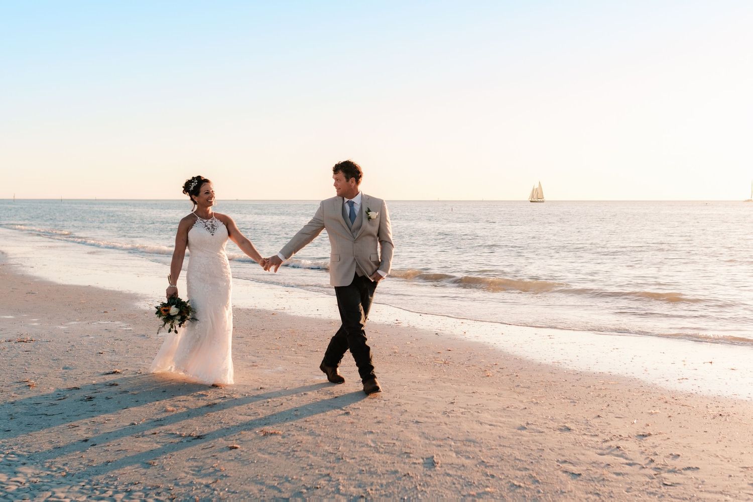 Bride and groom holding hands walking on sandy beach at sunset with sailboat on horizon.